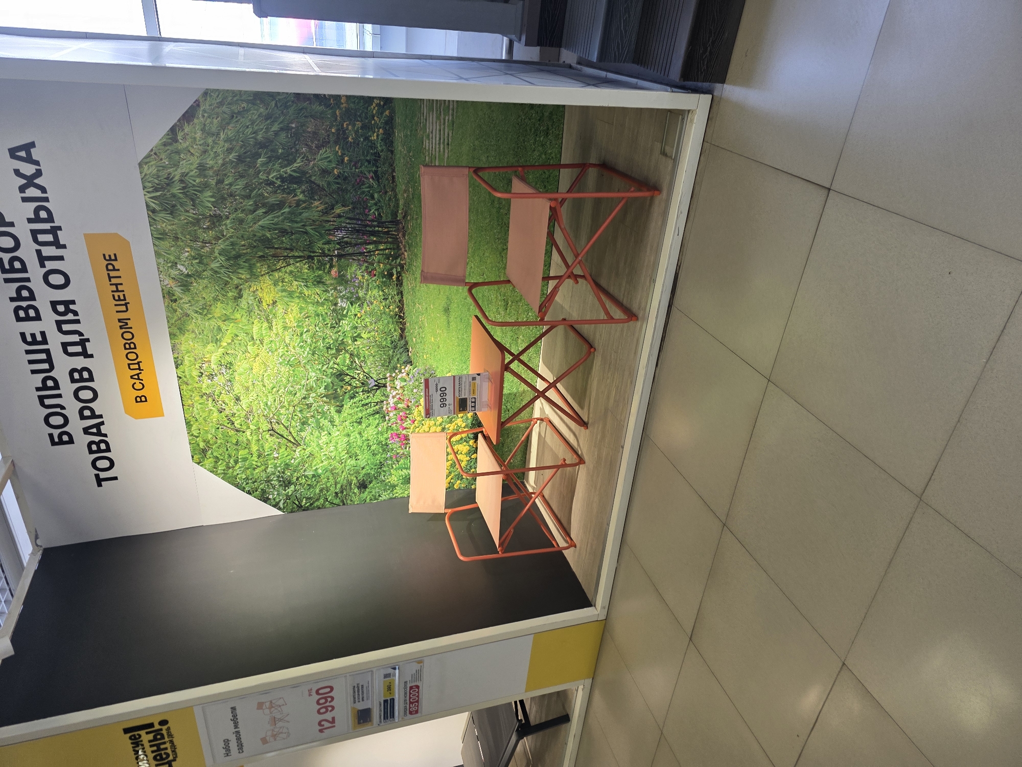 This is a medium-angle shot of a retail display featuring outdoor furniture. The display is set against a large photographic backdrop of a lush green garden with bamboo. In the foreground, there are two orange folding chairs and a small matching orange folding table. One chair is positioned to the left of the table, and the other is to the right. The chairs and table appear to be made of metal and fabric. To the left of the main display, a portion of a yellow and white sign is visible, which includes text and images of furniture. Below this sign, a price tag shows "12 990". Further to the left, a dark-colored lounge chair is partially visible. The flooring in front of the display is a light-colored tile. The display is enclosed by a white frame, and the overall scene suggests an indoor retail environment. The lighting is bright and even, consistent with store lighting. There are no people present in the image.