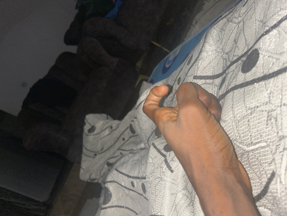 A close-up perspective shows a person's left hand clutching at a textured gray fabric with black line and circle patterns. The thumb is noticeably bent backward. The background is blurry, revealing parts of a dark brown sofa and a hint of a blue object. The lighting is dim, contributing to the shadowy appearance of the couch and surrounding area.