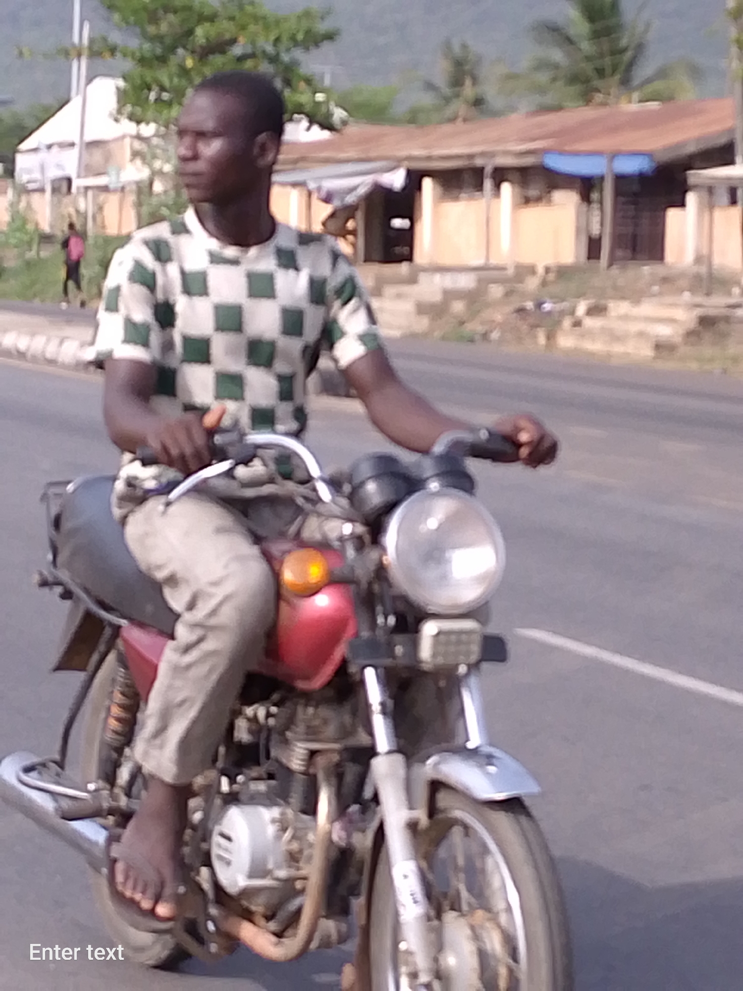 A man rides a motorcycle on a paved road in Adankolo, Nigeria. The man is wearing a checkered shirt and light-colored pants. He is barefoot. The motorcycle is red and silver. In the background are buildings and vegetation. A person is walking along the shoulder of the road in the background. The photo is taken in daylight.