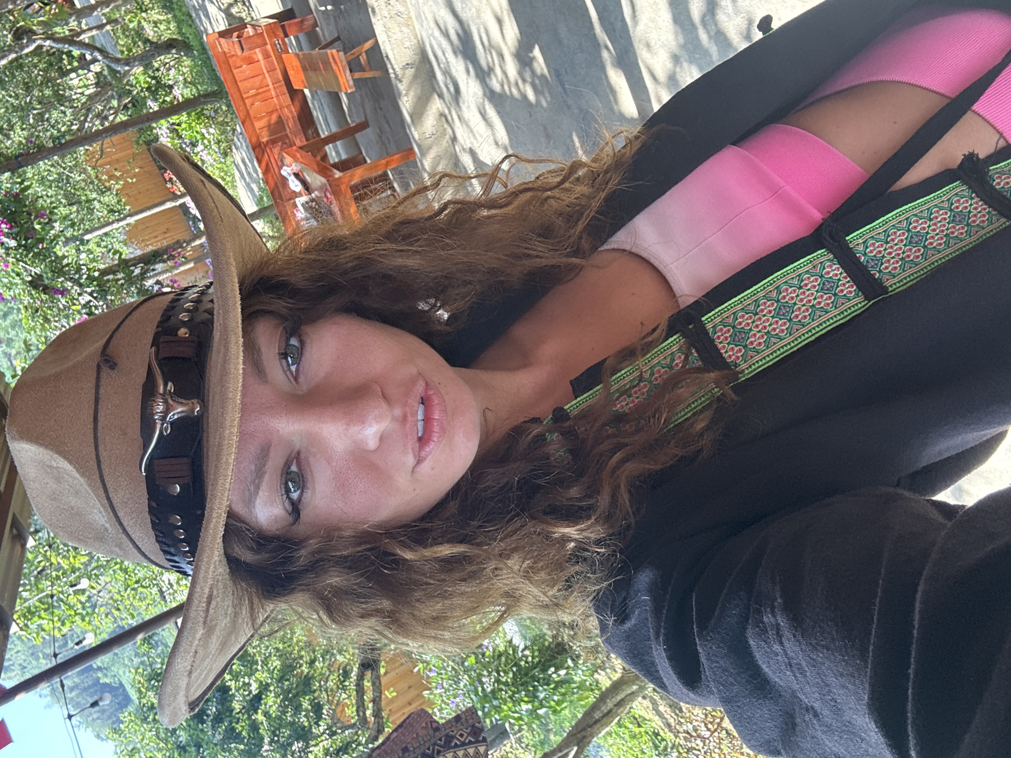 A woman wearing a cowboy hat poses outdoors, likely at an elevated location within the city of Bắc Hà, Vietnam. She has long, curly brown hair, light skin, and appears to be wearing pink clothing underneath a dark-colored outer garment with a decorative strap or trim. The hat is tan-colored and features a distinctive ornamental design on its band. The background includes visible vegetation, and partially visible buildings and furniture.