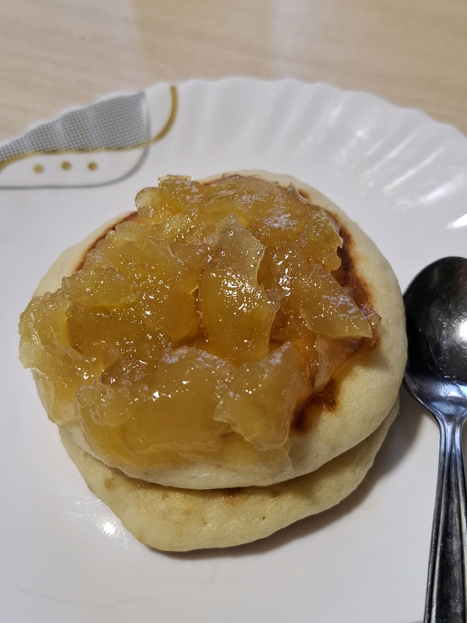 A close-up, overhead shot shows two small, golden-brown pancakes stacked on a white plate. The top pancake is generously covered with a glistening, amber-colored fruit compote, possibly apple or apricot, with visible chunks of fruit suspended in a thick, translucent syrup. A portion of a silver spoon is visible on the right side of the plate, positioned next to the pancakes. The plate has a subtle embossed pattern around its rim. The background is out of focus, appearing to be a light-colored wooden surface. There is no indication of the city of Tyumen, Russia, or any other geographical location. The image focuses solely on the food item.