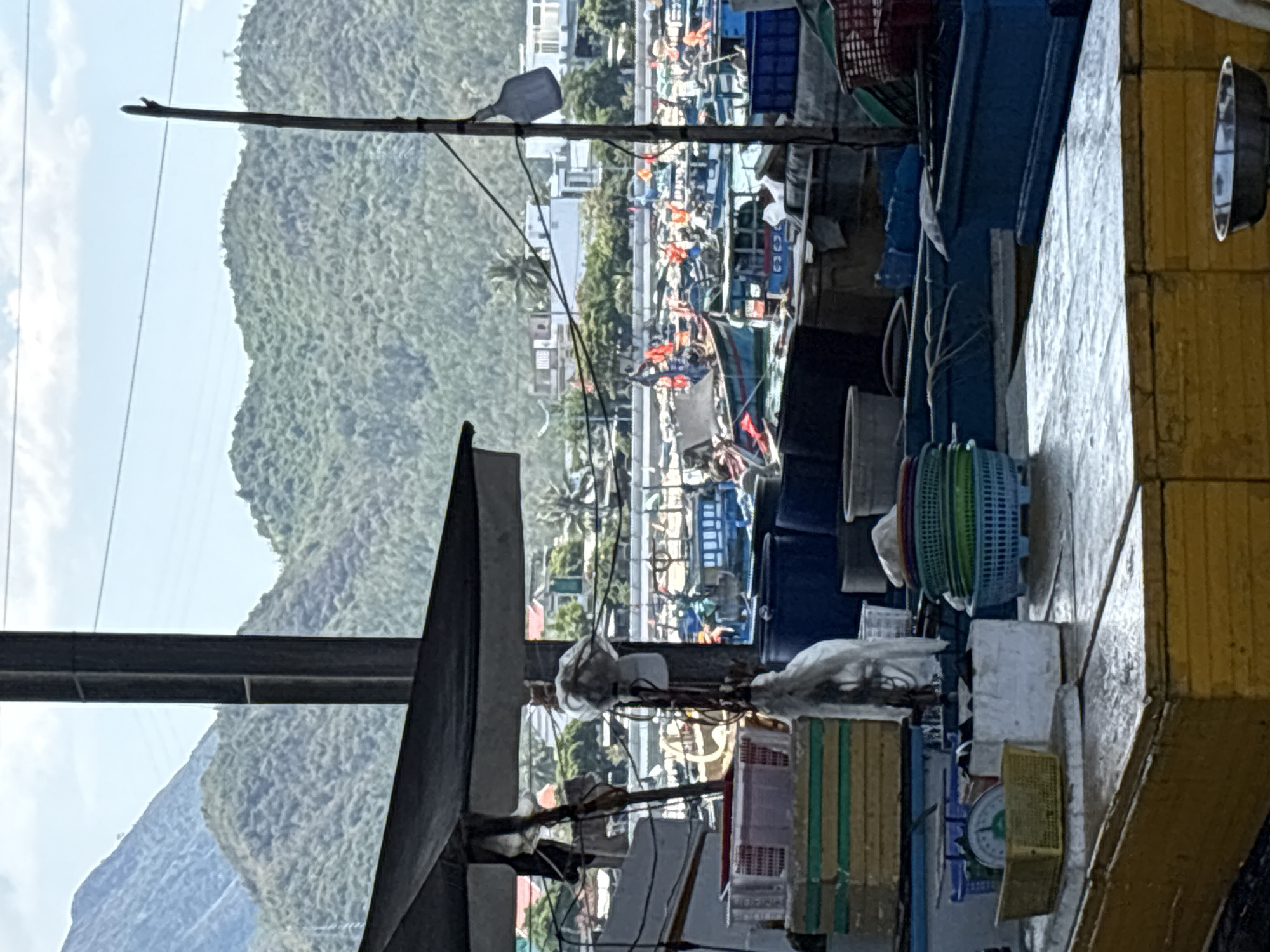 The image shows a cluttered scene in Vĩnh Lương, Vietnam, potentially near a harbor or fishing area. Numerous fishing boats are visible in the background, packed together and adorned with orange life vests. A hillside is also in the backdrop. In the foreground, there is a close-up view of boat elements, including a weathered yellow structure, ropes, baskets of varying colors, and other undefined equipment. The viewpoint suggests being either on a boat or near the docks, looking out toward the bay and village.
