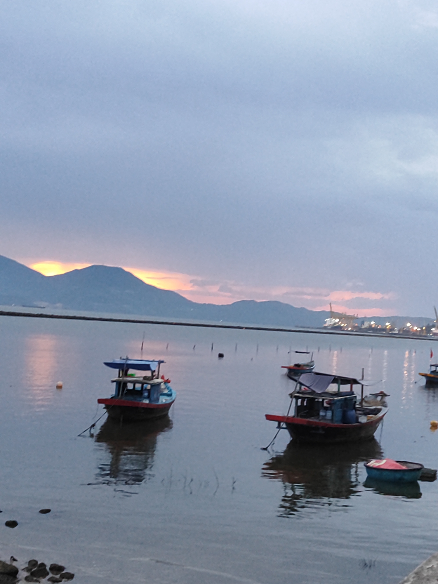 The image depicts a serene coastal scene at dusk or dawn, likely in the vicinity of Đà Nẵng, Vietnam. In the foreground, the calm water reflects the soft, diffused light of the sky. Several small fishing boats are moored or drifting on the water. One prominent boat in the mid-left has a blue cabin structure with a red hull. Another similar boat is positioned further to the right. A distinct round, colorful basket boat is also visible in the right foreground. In the background, a long, dark landmass, likely mountains, forms the horizon, with streaks of orange and yellow light peeking through the clouds at their peaks, indicating either sunrise or sunset. Beyond the water and mountains, a distant city skyline is faintly visible, dotted with lights, suggesting an urban area bordering the coast. The overall atmosphere is peaceful and tranquil.
