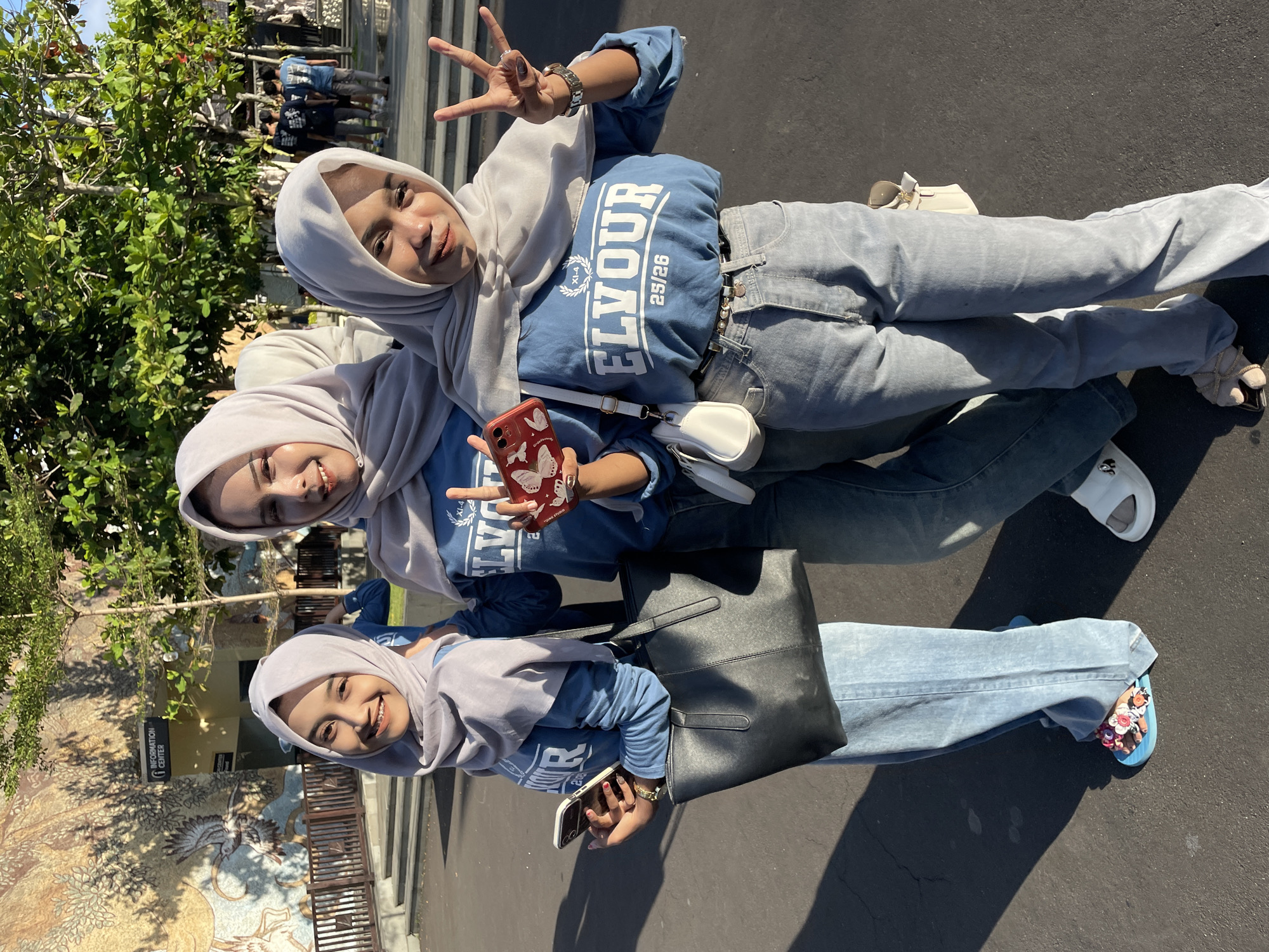 Three young women, likely friends, are captured in an outdoor setting, posing cheerfully for a photo. All three are wearing light grey hijabs and matching blue long-sleeved tops or sweatshirts with light blue jeans. They are smiling brightly, and two of them are making peace signs with their hands, conveying a sense of joy and friendship.
The woman at the front is holding a white mobile phone, while the middle woman holds a red mobile phone with a white butterfly design on its case. She also wears a watch and rings. The woman at the back holds a white shoulder bag with sunglasses resting on it. The middle woman has a black tote bag over her shoulder. Their footwear includes white sliders and light blue flip-flops adorned with colorful flowers.
The background reveals a sunny day with bright sunlight casting shadows. Lush green trees are visible on the left side of the frame. On the right, there's a detailed mosaic wall featuring a large bird and nature-inspired designs, alongside a railing. A building with an "INFORMATION CENTER" sign and an arrow is partially visible behind the women. Further in the distance, a group of people can be seen. The ground they are standing on appears to be a paved path or road.
Visible text includes "ELYOUR 25/26" and "XL-4" on the blue tops. The "INFORMATION CENTER" sign is also clearly readable. The overall scene suggests a pleasant outing at a public location, possibly a tourist attraction or park in Kuta Selatan, Indonesia.