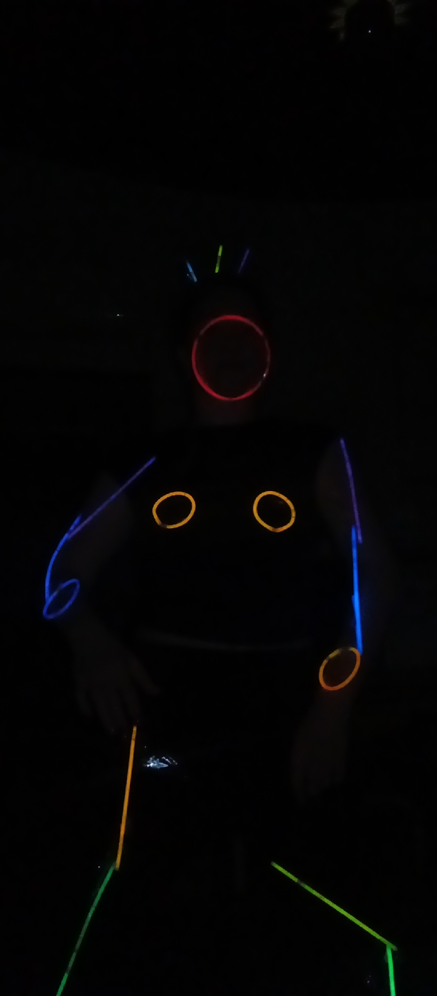 A person in an unknown location is wearing a black outfit with glow sticks attached to create a stick figure effect. There are glow stick rings on the head, arms, and chest, and glow sticks forming lines on the limbs.
