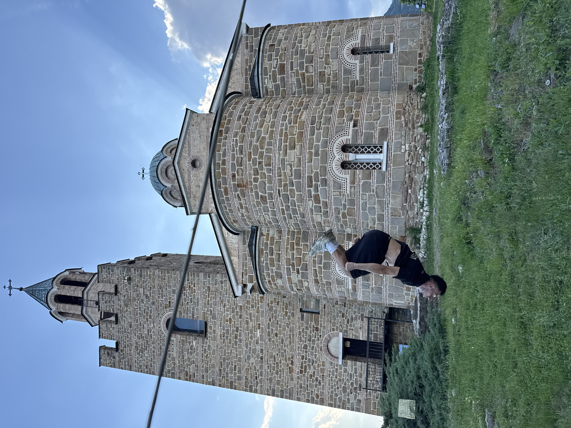A man in black clothing is performing a backflip on a grassy area next to a stone building. The building appears to be a historic church or monastery, judging by its stone construction, arches, windows, and cross-topped towers. The sky is blue with some clouds and sun rays. The building's architecture suggests a religious or historic structure, likely located in Europe. Based on the architecture, it is the Holy Forty Martyrs Church in Veliko Tarnovo, Bulgaria.