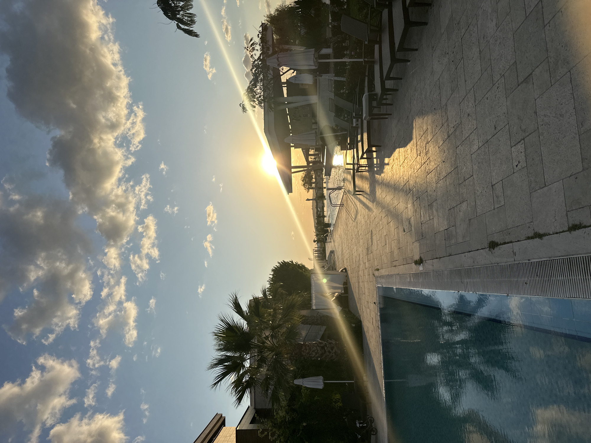 The image depicts an outdoor scene at what appears to be a resort or hotel complex near the city of Sozopol, Bulgaria. The time of day is sunset, as evidenced by the low sun angle and the golden light casting long shadows.
In the foreground, a swimming pool with blue water is visible on the left, its surface reflecting the sky and surrounding palm trees. The pool is bordered by a tiled walkway. Further along the walkway, lounge chairs and umbrellas are arranged near a covered bar area, suggesting a leisure and relaxation setting. Palm trees and other vegetation are scattered throughout the scene, adding to the tropical or Mediterranean ambiance. The sky is a mix of blue with scattered clouds, some illuminated by the setting sun. The architecture visible on the left suggests modern buildings associated with the resort. The overall impression is one of a tranquil evening at a well-appointed outdoor venue.