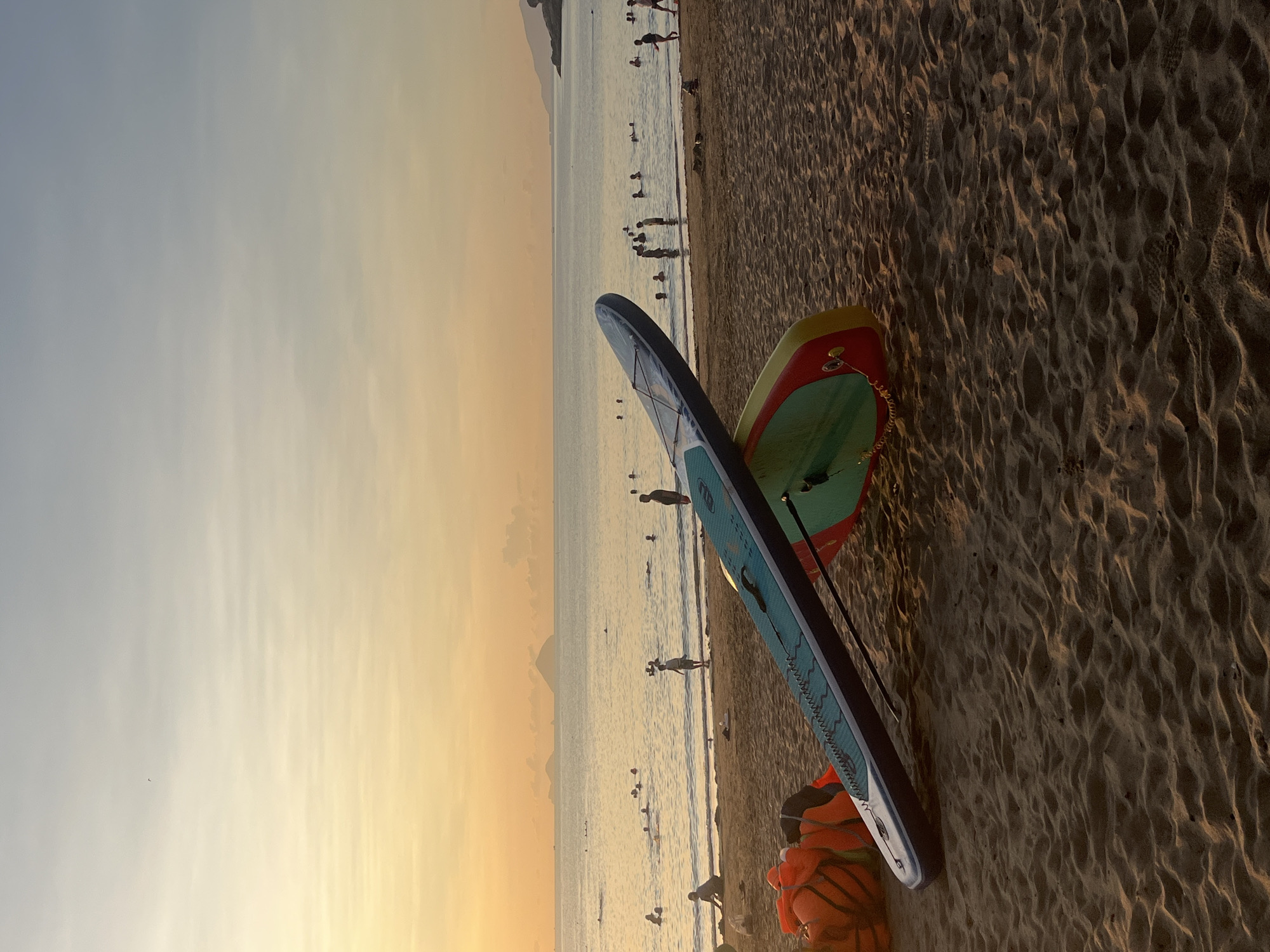 Two paddleboards are propped up against each other on a sandy beach in Bac Nha Trang Ward, Vietnam. One is primarily blue with white and turquoise accents, and the other is green with red and yellow. Orange life jackets are bunched up near the base of the paddleboards. People are swimming and standing in the water in the background. The sky is an orange and yellow gradient, suggesting sunset.