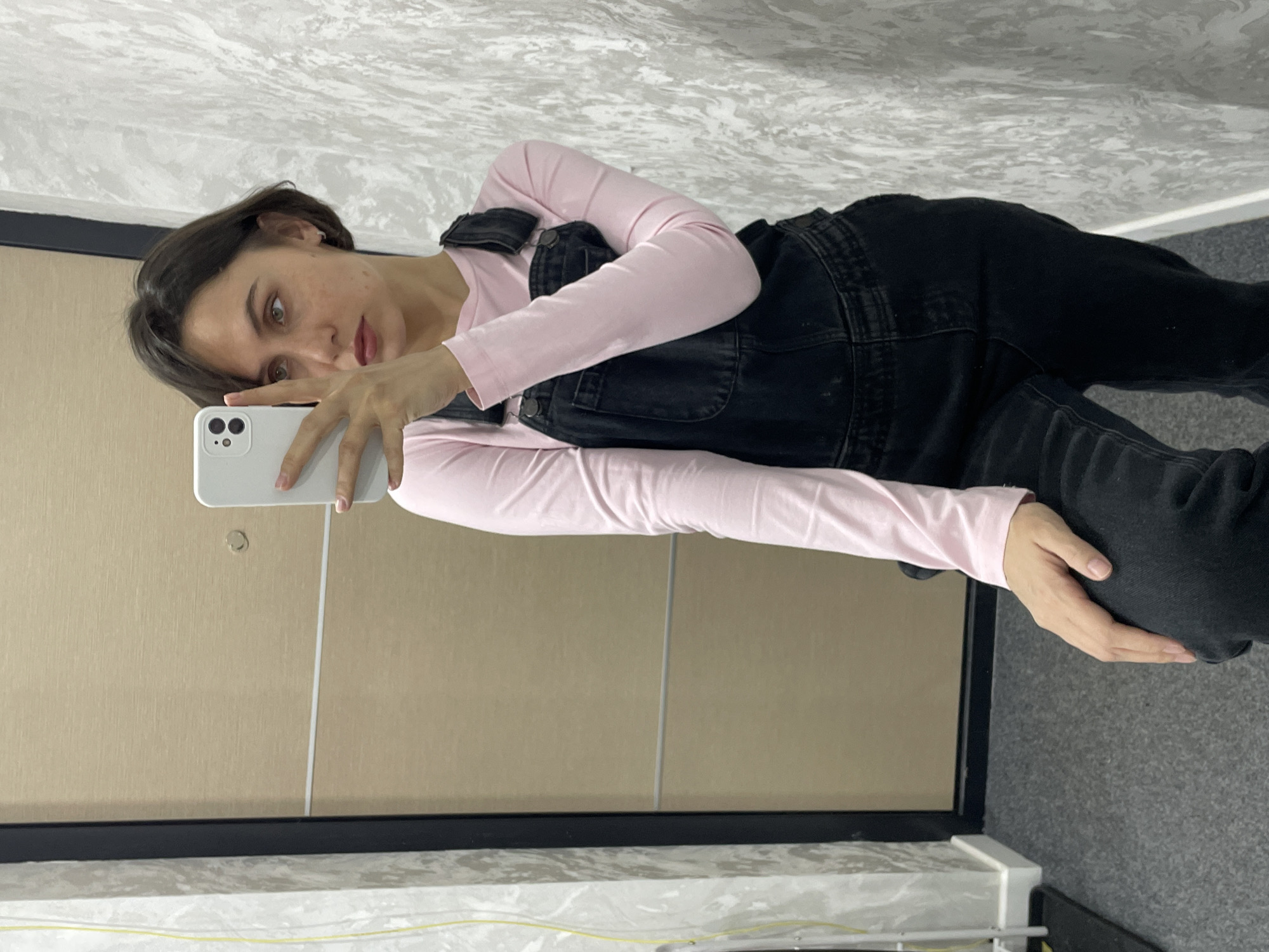 A young woman is captured in a mirror selfie, looking directly towards the camera with a neutral to slightly pensive expression. She has short, dark hair, and light-colored eyes (appearing greenish or hazel), with visible natural blemishes or freckles on her cheek. She holds a white smartphone, likely an iPhone, in her right hand to take the picture, showcasing light pink or nude polished fingernails.
She is dressed in a light pink long-sleeved top, layered under dark, possibly black, denim overalls. The overalls feature visible seams and buttons.
The setting appears to be an indoor space, possibly a hallway or room in Izhevsk, Russia. The background features a light beige or cream-colored wall with a smooth finish. A vertical white strip, likely a door frame or molding, is visible on the wall. Above this, a distinct grey and white textured pattern, resembling marble or concrete, covers the upper wall or ceiling, separated by a dark horizontal trim. The floor is covered with a grey carpet. The lighting is bright and even, typical of an indoor environment. No text is visible in the image.