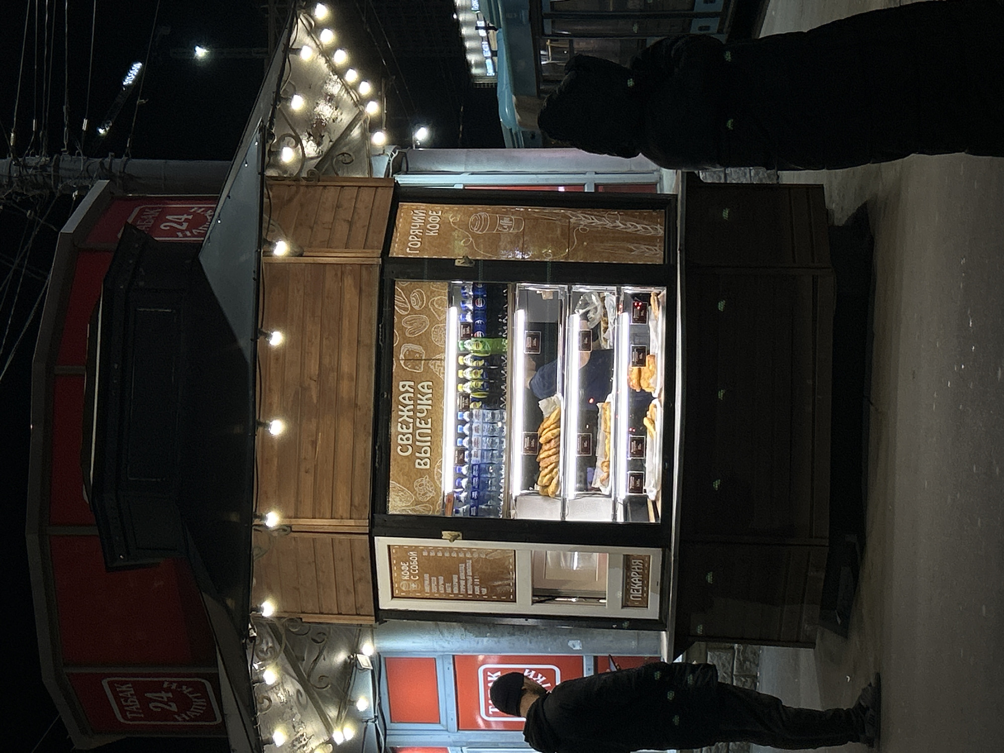 The image depicts an urban night scene, likely in Saint Petersburg, Russia, featuring a brightly lit street kiosk. Two individuals, dressed in dark winter clothing, are visible near the kiosk; one stands in front, and the other further back. The kiosk itself is adorned with warm string lights and has wooden paneling and glass display cases.
Inside the kiosk's display, various baked goods such as pastries and possibly sausages in dough are visible, along with rows of bottled drinks in a refrigerated section. The kiosk appears to be a bakery and coffee stand.
Several signs with Cyrillic text provide details about the offerings. A large sign on the kiosk reads "СВЕЖАЯ ВЫПЕЧКА" (Fresh Baked Goods) and "ГОРЯЧИЙ КОФЕ" (Hot Coffee), accompanied by illustrations of a coffee cup and wheat. Another sign on the lower part of the kiosk lists coffee types under "КОФЕ С СОБОЙ" (Coffee To Go), including Latte, Cappuccino, Americano, and Espresso. The word "ПЕКАРНЯ" (Bakery) is also clearly visible. To the left of the main kiosk, a red sign advertises "ТАБАК" (Tobacco) and "24", indicating a 24-hour tobacco shop nearby.
The scene is entirely dark except for the artificial lighting from the kiosk and distant streetlights, suggesting late evening or night. The presence of people and the open kiosk indicate active business.