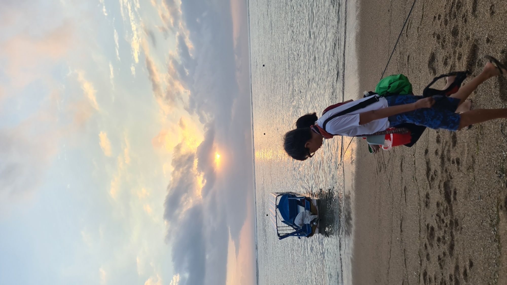 An outdoor, eye-level, full shot shows two people walking along the beach at Sanur, Denpasar, Indonesia, during sunset. The sky is a gradient of pale blue and orange, with scattered clouds. The sun is partially obscured by clouds, casting a golden reflection on the ocean's surface. The water is calm, with small waves gently lapping the shore. A blue and white boat with a canopy is anchored near the shore. The sand is light brown and speckled with small pebbles, with visible footprints.
The person in the foreground, viewed from the side, is wearing glasses, a white t-shirt, blue patterned shorts, and flip-flops. They are carrying a red bucket in their left hand and a green bag slung over their right shoulder. A second person, partially visible behind the first, appears to have dark hair and is wearing a red scarf. They are walking in the same direction. The overall scene evokes a peaceful and serene atmosphere.