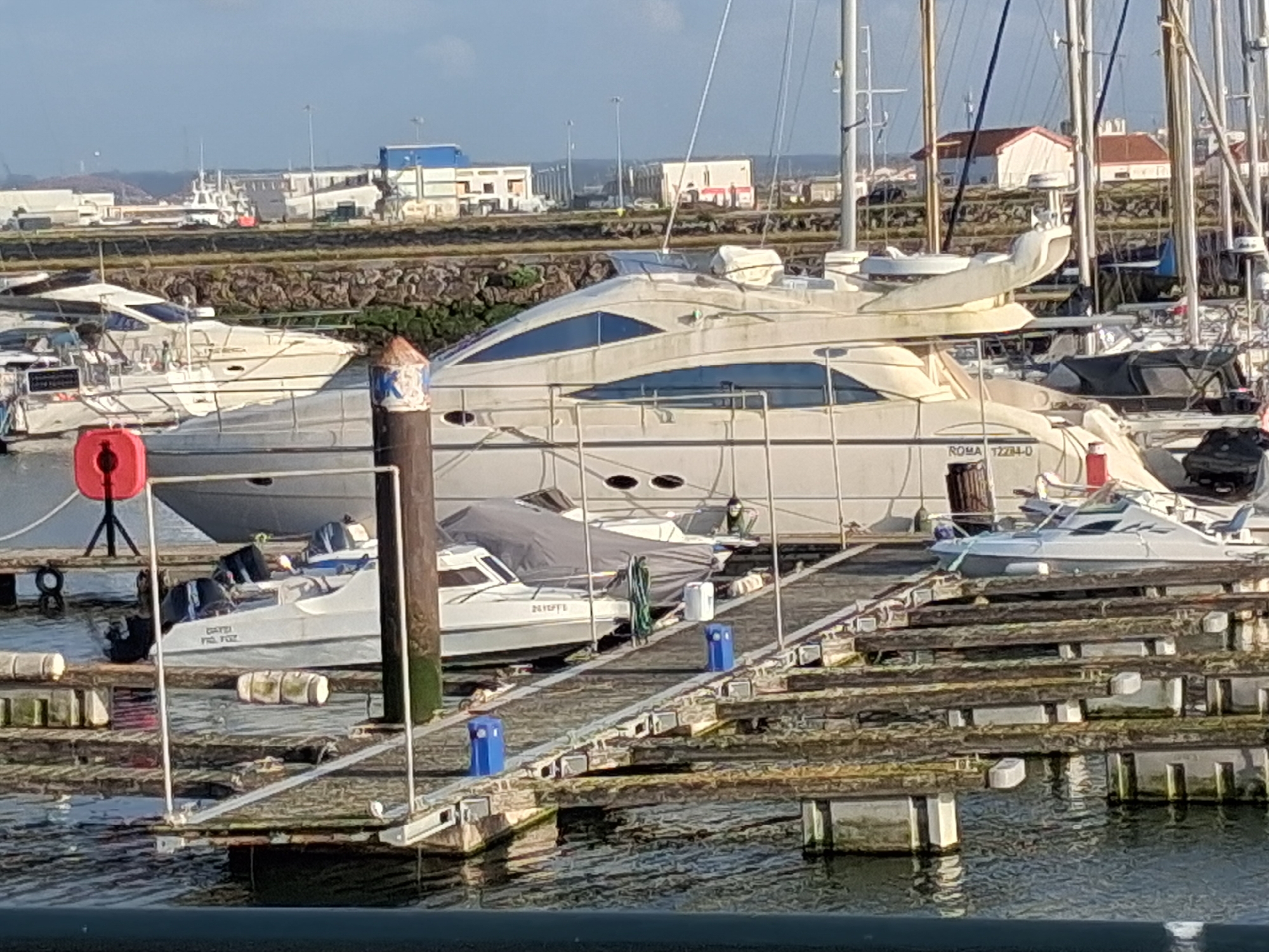 The image captures a marina in Figueira da Foz, Portugal, featuring a large, off-white yacht prominently positioned among other boats. The yacht displays the registration "ROMA 12284-0". Various smaller boats are docked nearby, some covered. Wooden docks, weathered and multi-tiered, extend into the water. In the background, buildings line the waterfront, with some appearing as white structures with blue and red accents. Masts and rigging of other sailboats are visible in the periphery. The water reflects the daylight, and the overall scene conveys a serene harbor setting.