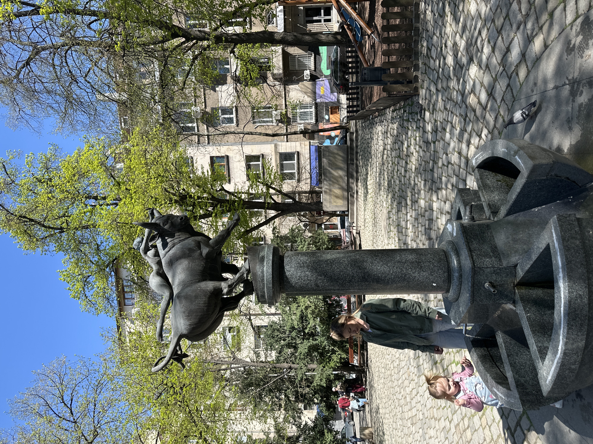 The image shows a street scene in Sofia, Bulgaria, featuring a statue of Europa and the bull in Borisova Gradina Park. A woman and a young child are next to a water fountain. The statue is mounted on a dark grey cylindrical pedestal. The environment includes trees with green foliage, a cobblestone ground, and a multi-story building in the background. A pigeon stands near the fountain.