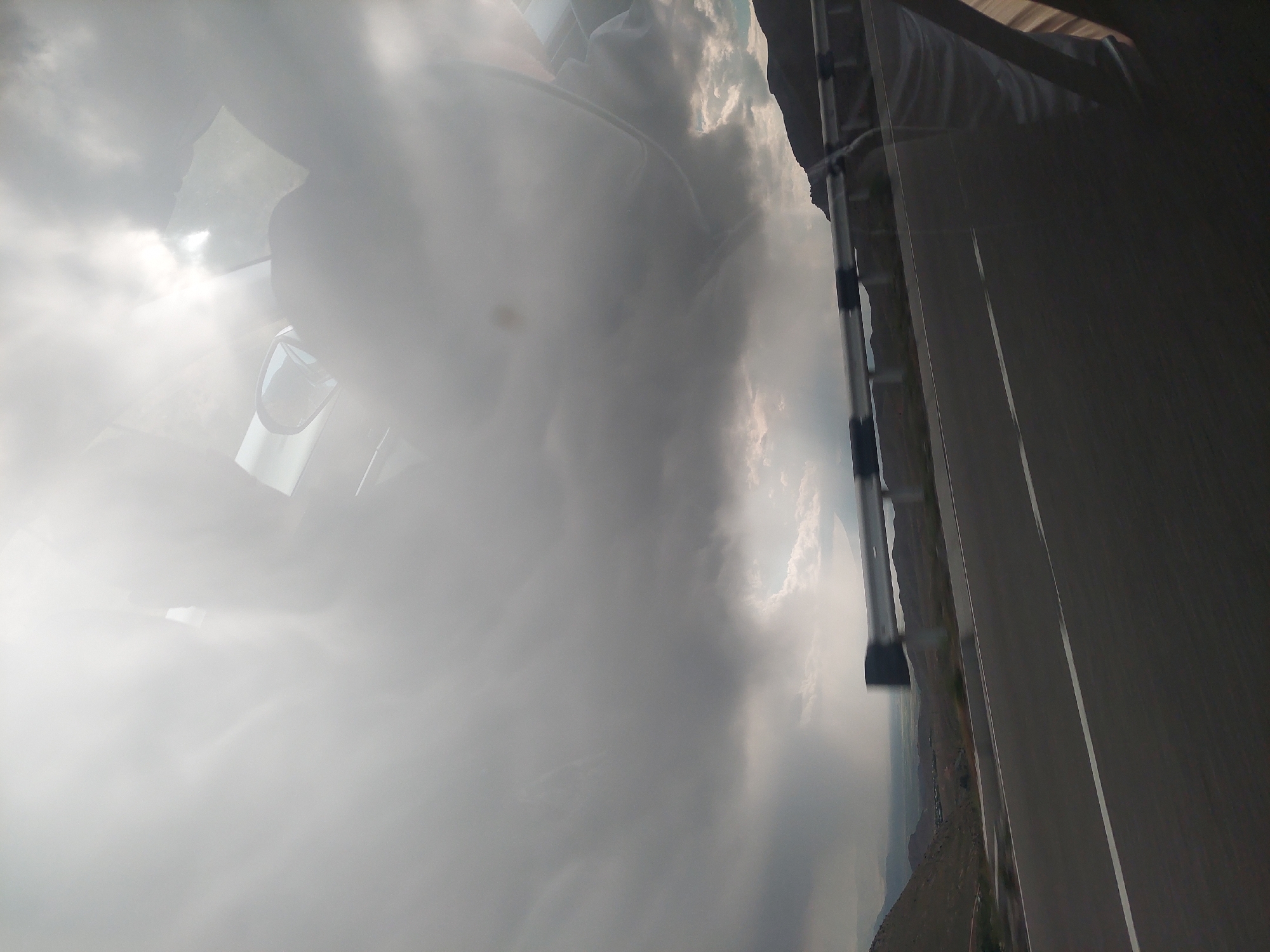 The image captures a dramatic outdoor scene, likely taken from inside or very close to a moving vehicle. The dominant feature is a heavily clouded sky, with thick grey clouds interspersed with areas where brilliant sunlight breaks through, creating strong highlights and lens flares, particularly on the left side.
In the upper-left portion, reflections are visible on what appears to be a car window or sunroof. These reflections include distorted shapes of the clouds and possibly elements of the car's interior, such as a sun visor or dashboard.
To the right, a paved road with a clear white line extends into the distance. Alongside the road, a rugged, mountainous, or hilly landscape is visible, suggesting a dry or arid environment. A metallic structure, possibly a roof rack or part of the vehicle's exterior, is present along the right edge of the frame.
The time of day appears to be daytime, characterized by intense sunlight despite the heavy cloud cover. The weather is dynamic, with bright, breaking sun through an otherwise overcast sky. The location context of Tigranashen, Armenia, aligns with the visible mountainous terrain. No people or visible text are present in the image. The scene evokes a sense of travel and observation of the vast, natural landscape under striking weather conditions.