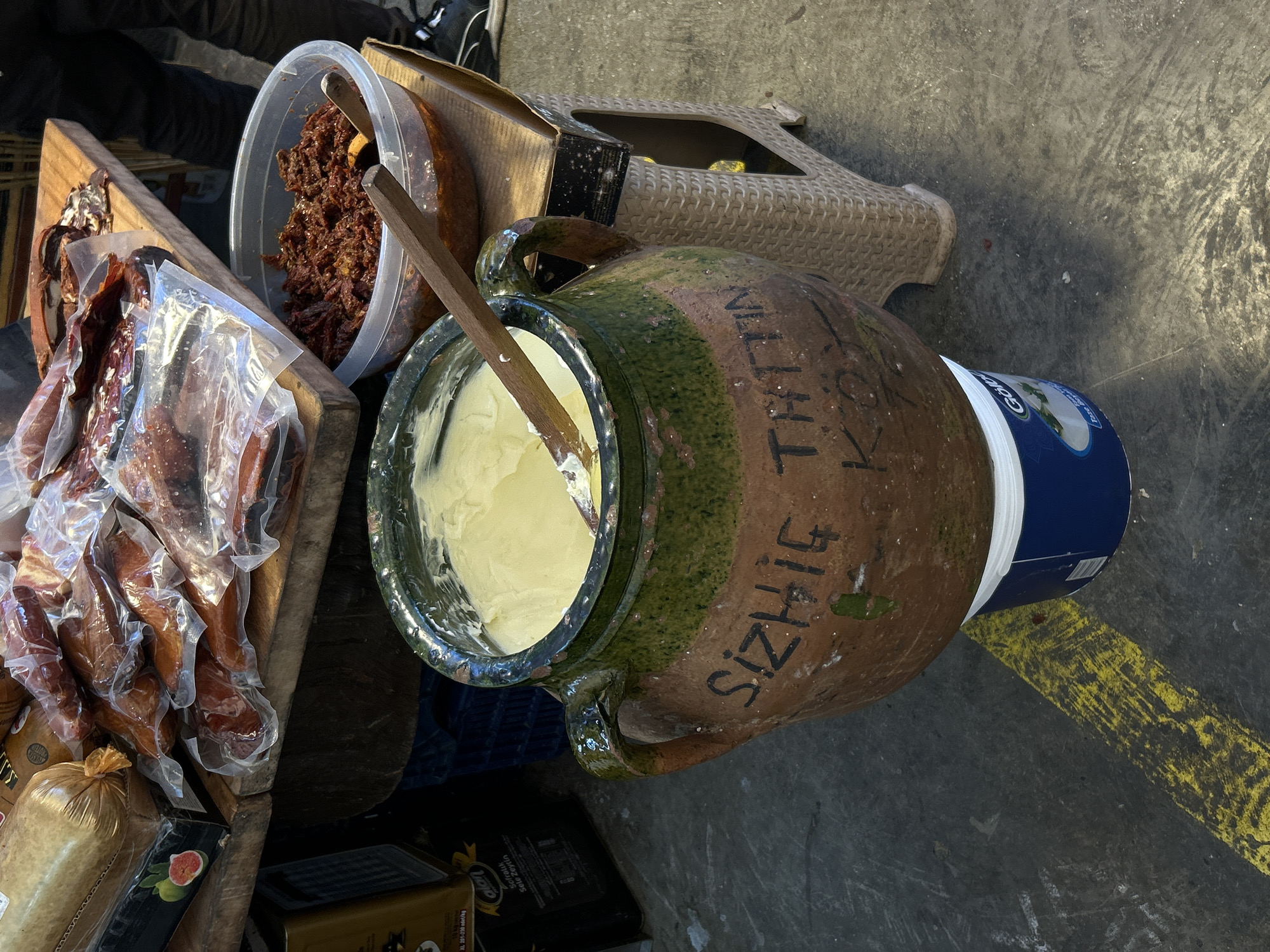 This image shows a market scene, likely in Alanya, Turkey, based on the Turkish writing on the ceramic jar. The jar, containing a yellowish cream-like substance and a wooden spoon, sits atop a blue and white container. To the left, there is a wooden structure holding packages of meat in plastic wrap, a clear plastic container filled with a dark red, pasty substance, and other various goods. The background includes a beige plastic stool. The scene is set on a gray surface, possibly a street or market floor, marked with a yellow line. Part of a person's legs and footwear are visible in the top left corner.