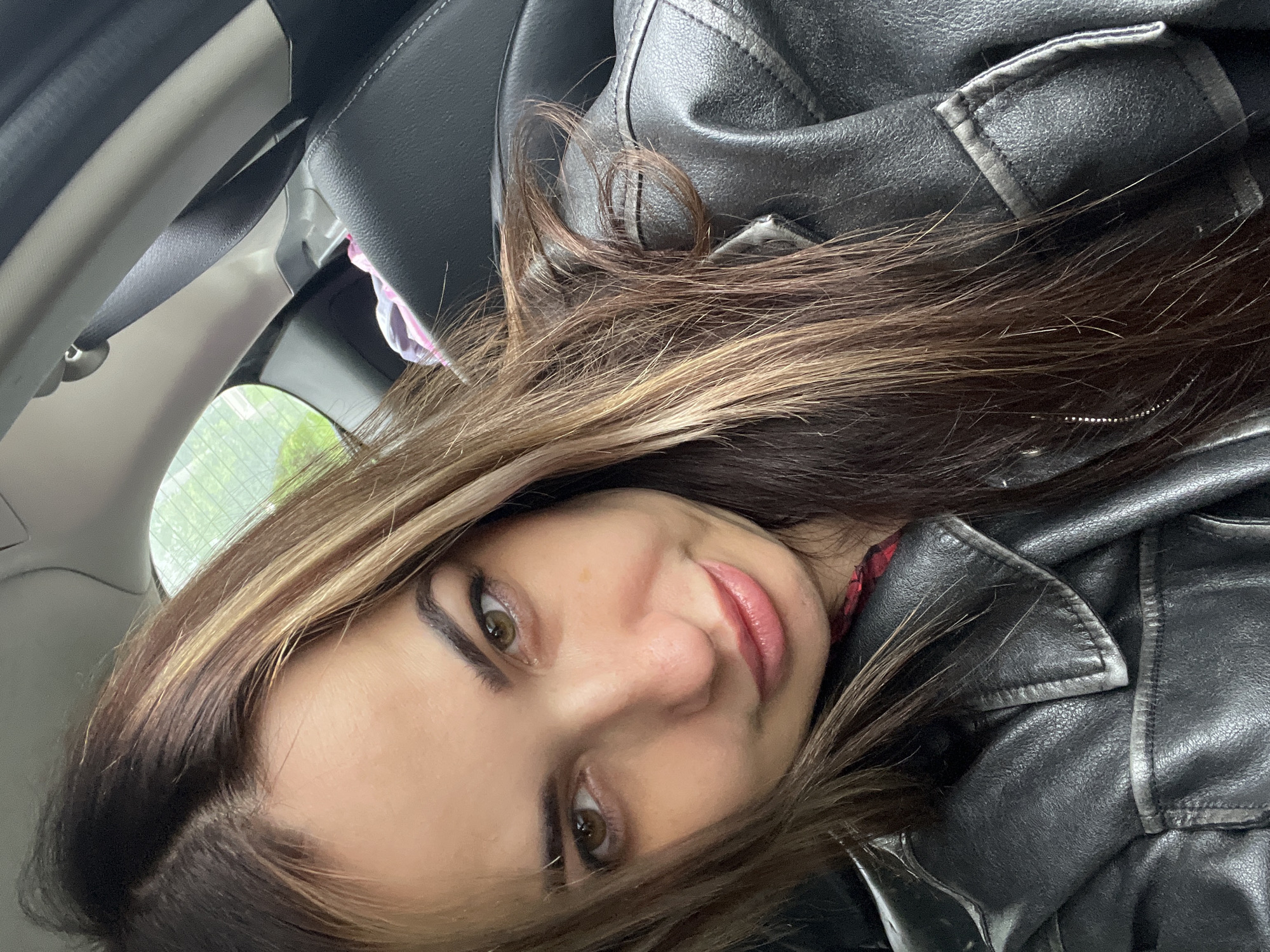 A female subject is depicted in a self-portrait taken inside a vehicle in the city of Unknown, Unknown. The subject, positioned centrally, has long, brown hair with lighter blonde or light brown highlights, and visible dark root regrowth. Her hair is parted on her left side (viewer's right). She has brown eyes, dark, well-groomed eyebrows, a straight nose, and full lips with a pinkish-red lip color, displaying a subtle smirk. Her complexion is fair to medium, with minor blemishes or freckles visible on her right cheek. She is wearing a dark, possibly black or dark grey, leather or faux-leather jacket with a prominent collar and visible stitching. A glimpse of a red and black plaid garment is visible beneath the jacket. The vehicle's interior features a dark car seat with light contrast stitching, a black seatbelt fastened to a light grey B-pillar, and a light-colored headliner. Through a rear window on the upper right, blurred green foliage is visible, indicating an outdoor setting. A pink and white patterned item is partially visible on the rear seat.