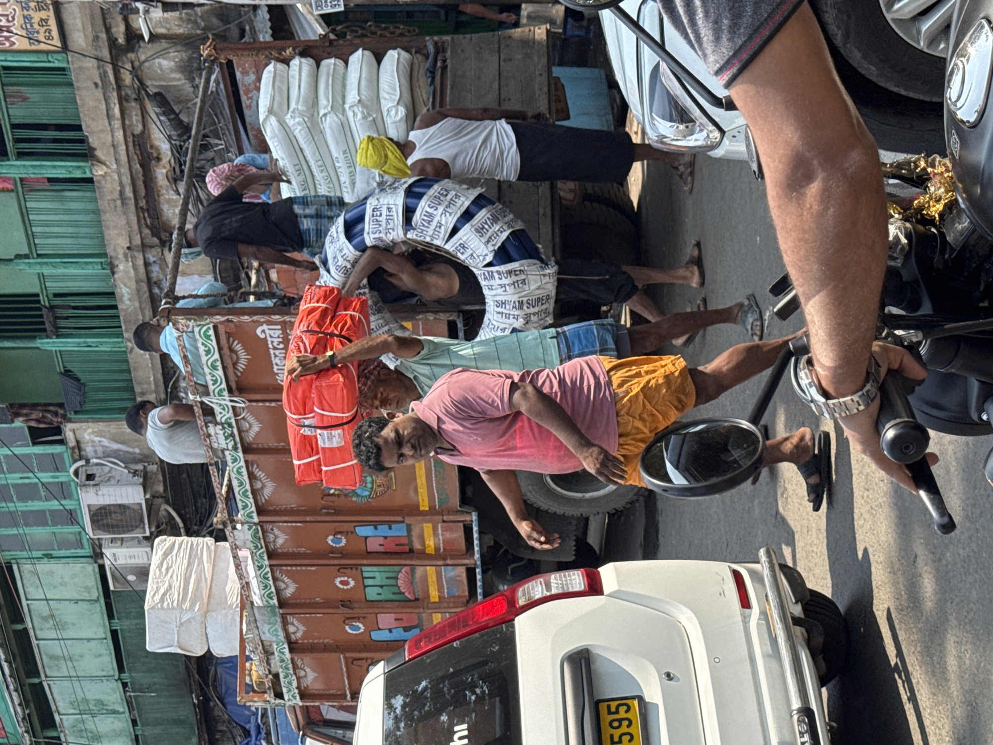 This image captures a bustling street scene in a commercial district of Kolkata, India, under clear daylight conditions.
In the center, multiple male laborers are actively engaged in the manual loading and unloading of goods around a large, ornately painted brown cargo truck (lorry). The truck's body features decorative white and red patterns and Bengali script. It is laden with numerous white sacks, several of which are marked "MAIZE STARCH 50 KG. NET," and other packaged items.
To the immediate left of the truck, a man in a pink polo shirt, lightly stained with red powder, and orange shorts walks towards the viewer, looking directly at the camera. His bare feet are visible, wearing black flip-flops. Behind him, another man carries multiple red-wrapped bundles stacked on his head. Further right, a laborer shoulders a large, curved bundle of blue pipes, prominently wrapped in white plastic with "SHYAM SUPER" text. Near the rear of the truck, another man in a white tank top and dark lungi, with a yellow cloth wrapped around his head, stands beside a stack of white sacks. Several other men are visible on and around the truck, handling cargo.
In the foreground, the rear of a white Maruti Suzuki Wagon R car is visible on the left, displaying a partial yellow license plate "595." The right foreground shows the handlebar, mirror, and the forearm of a person riding a motorcycle, indicating the image was taken from a two-wheeler.
The background features multi-story, aged buildings with green-painted windows and balconies, some equipped with exterior air conditioning units. The street is asphalt, and shadows suggest mid-morning or afternoon sun. The presence of Bengali script on the truck and goods, alongside the characteristic urban architecture and vehicle types, confirms the location as Kolkata.