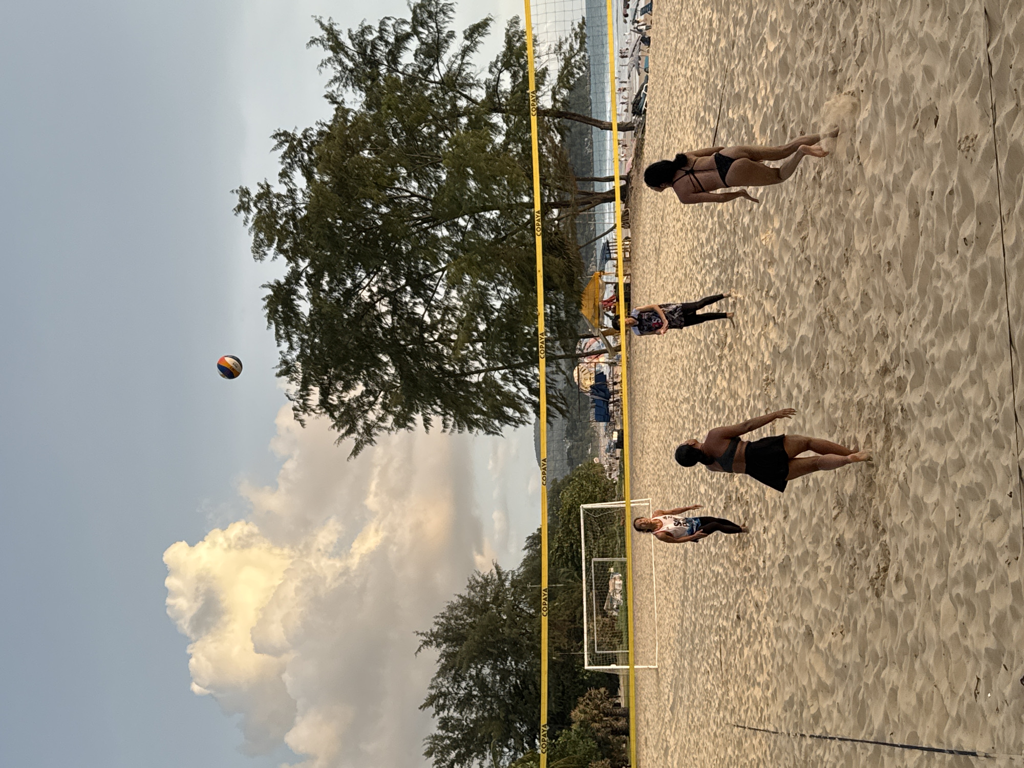 In Karon, Thailand, a beach volleyball game is underway on a sandy court. Four individuals are visible on the court, engaged in play. In the foreground, two individuals are positioned on the right side of the court, facing away from the camera. One player, wearing a black bikini top and a black skirt, is in motion, kicking up sand as they run. The other player, slightly further back and to the left, is facing forward and appears to be watching the game. On the left side of the court, two more individuals are present. One player, wearing a white tank top and black leggings, is looking upwards. Another player, wearing a patterned top and a headscarf, is facing towards the net. A volleyball is in the air above the players, indicating an active game. The volleyball court is marked by a yellow net, with white lines on the sand. In the background, a soccer goal is visible behind the net. Beyond the court, trees line the beach, and in the distance, the ocean and a populated coastline can be seen. The sky is a pale blue with scattered clouds, suggesting it is late afternoon or early evening.