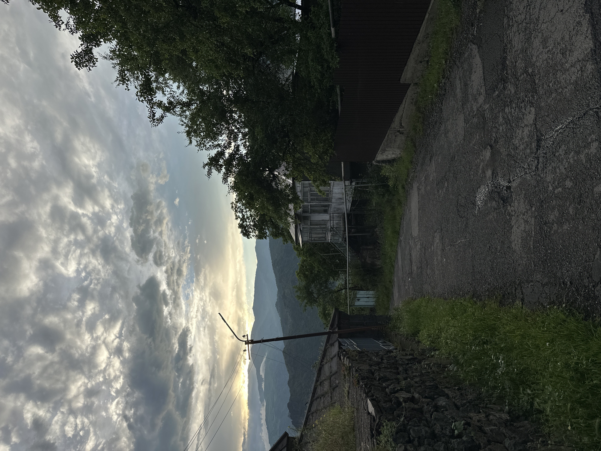This image depicts a rural road in the village of Haghpat, Armenia, under a cloudy sky. A narrow, asphalt road curves uphill, flanked by a rough stone wall on the left and a dark wooden fence on the right. Overgrown green grass and weeds sprout along the base of the wall and fence.
In the background, a two-story house with a weathered white exterior and a porch with windows is visible, partially obscured by dense green trees. Power lines are strung across the scene, supported by a utility pole. Beyond the immediate vicinity, layers of rolling, mist-covered hills and mountains stretch into the distance, with the sun setting or rising behind them, casting a soft, diffused light through the clouds. The overall atmosphere is tranquil and suggests a remote, natural setting. No people or animals are present in the frame.