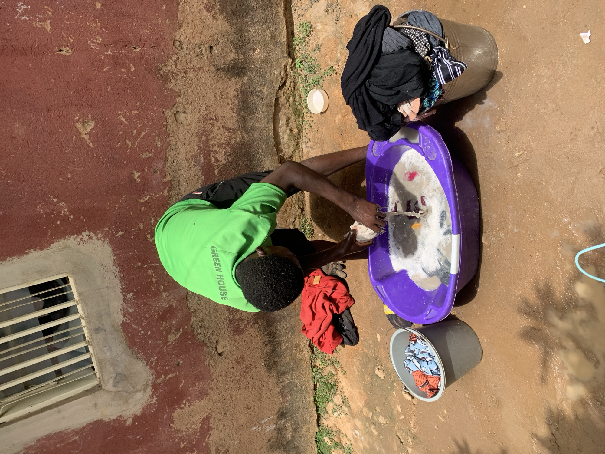 A person is hand-washing clothes outdoors in Jos, Nigeria. The individual is wearing a bright green t-shirt with the words "GREEN HOUSE" visible on the left sleeve. They are kneeling next to a purple plastic tub filled with soapy water and clothes. The person is actively washing clothes in the tub. A dark gray bucket filled with dirty laundry is next to the purple tub. A large metal bucket with dirty clothes overflowing is visible. Red and black clothing is on the ground near the individual. A white plastic lid is nearby. The scene is taking place outdoors, near a red wall with a barred window. The ground is dirt.
