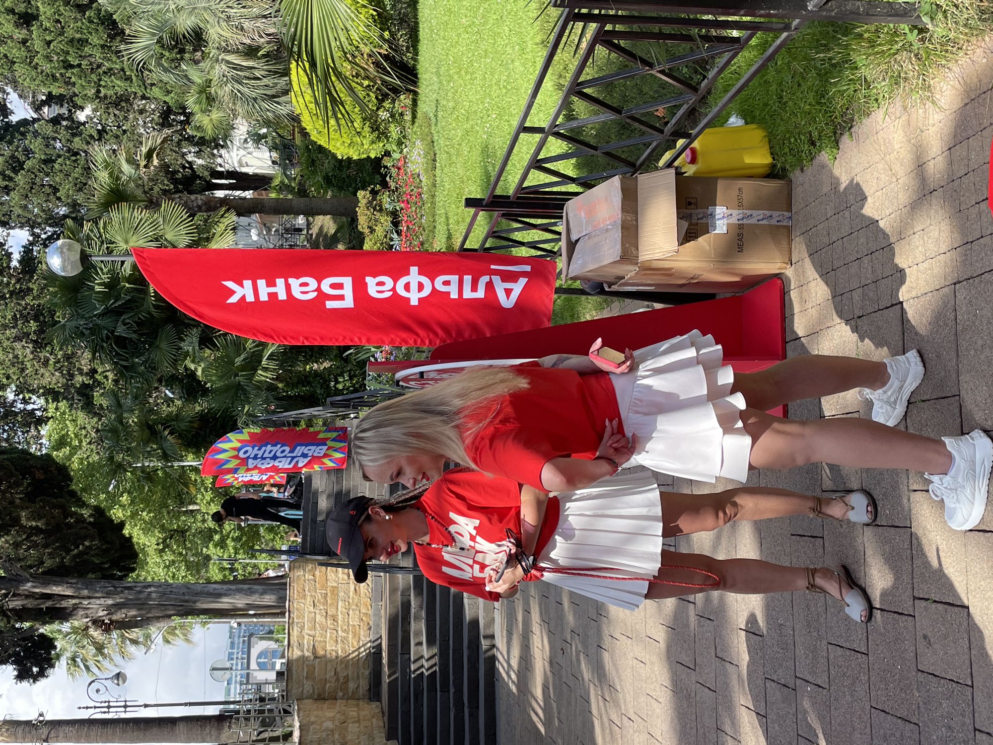 Two young women, dressed in red shirts and white pleated skirts, are standing outdoors on a sunny day, likely in Sochi, Russia, given the visual cues of palm trees and lush greenery. They appear to be participating in an event, possibly a promotional activity. One woman, wearing a black baseball cap, is looking down at something she holds in her hands, while the other, with blonde hair, leans in as if in conversation or observation. They are partially visible from the waist down.
In the background, a large red banner with white text is displayed vertically. The text, written in Russian, reads "Альфа Банк" which translates to "Alfa-Bank". Another smaller banner with colorful zigzag patterns and Russian text, "ВЫГОДНО" (meaning "profitable"), is also visible. Several cardboard boxes and a yellow plastic container are stacked near a metal fence. The setting suggests an outdoor event or promotion, possibly near a park or public space. The overall mood seems positive and energetic, with the women appearing engaged in their activity. The lighting indicates it is daytime, likely mid-morning to afternoon, given the bright sunlight casting shadows.