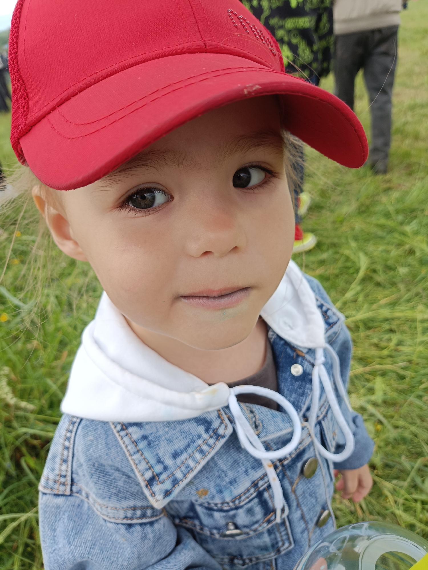 A young child, appearing to be a toddler or preschooler, is depicted in a medium close-up, gazing directly at the viewer. The child wears a red baseball cap with a slightly curved brim, a blue denim jacket over a white hooded sweatshirt, and a dark-colored top underneath. White drawstrings from the sweatshirt are visible and prominent in the foreground. A faint green stain is present on the child's lower lip. The child's right hand, partially obscured, holds a clear, spherical or bulbous plastic object in the bottom right corner of the frame. The background consists of blurred, unmown green grass, indicating an outdoor setting in the city of Unknown, Unknown. Partially visible adult figures are present in the distant, blurred background. The lighting appears soft and even, without harsh shadows.