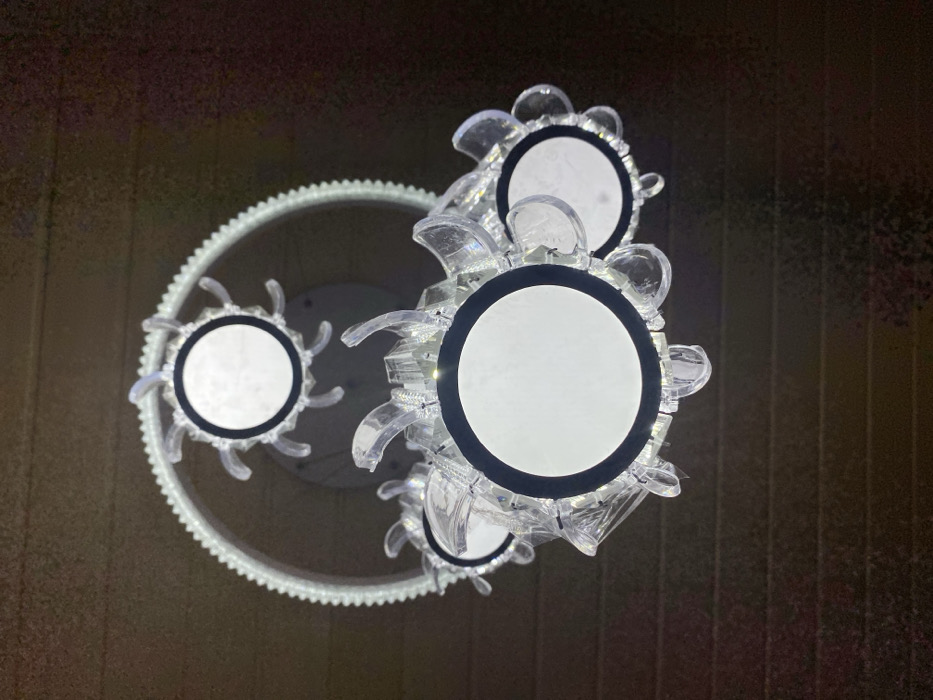 This image displays a modern, illuminated multi-component light fixture suspended from a dark, horizontally textured ceiling. The fixture, located in Tema, Ghana, features a prominent outer ring composed of numerous small, white, cog-like elements. Within this ring, a smaller circular light source with a white diffuser and a black rim is visible, surrounded by clear, jagged, or petal-shaped decorative elements. Below and partially overlapping this upper structure is a cluster of four additional circular light sources. Each of these also has a white diffuser, a black rim, and is encircled by clear, translucent decorative pieces that resemble crystalline petals or sharp facets. All visible light sources are actively illuminated, emitting a bright white light against the dark, possibly wooden or paneled, ceiling. No people, specific actions, or interactions are present in the scene.