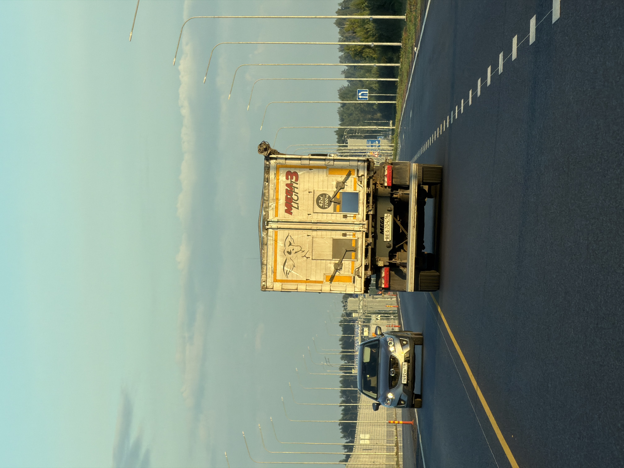 In Kaipy, Russia, a truck and a car are traveling on a highway. The truck, a large white vehicle with "MEGA LIGHT 3" written on its side, is in the right lane. A car, a silver SUV, is in the left lane behind the truck. The highway is flanked by streetlights and trees. The sky is blue with some clouds. The road has yellow and white lines.