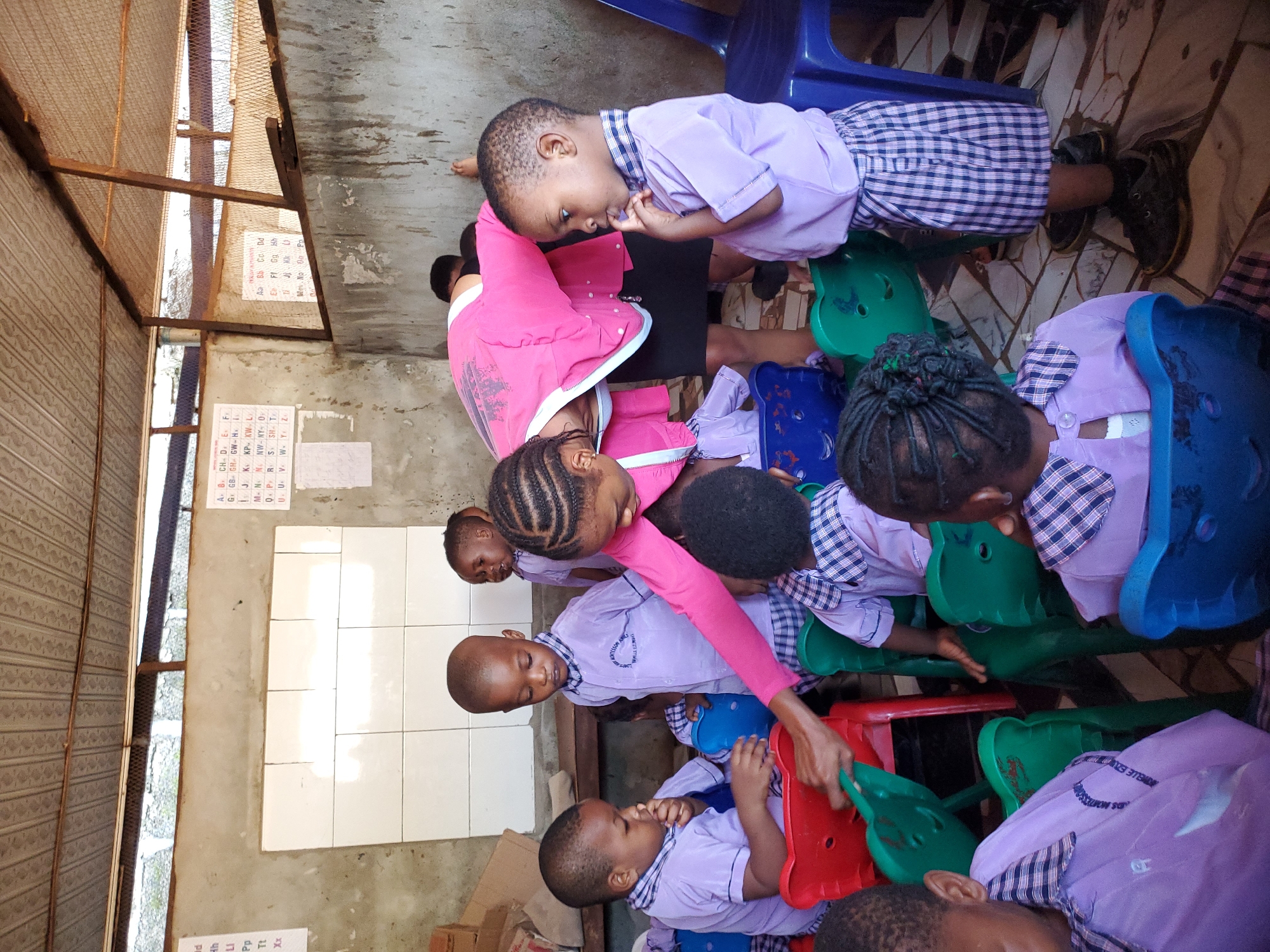 A slightly overhead, rotated shot captures a group of young children and an adult in what appears to be a modest classroom or learning space in Ukwu-Abwa, Nigeria. The scene shows several children, likely preschoolers or early primary students, dressed in purple uniforms with checkered collars and shorts/skirts. They are seated on small, brightly colored plastic chairs (red, green, and blue), focused on an activity or interaction.
An adult, possibly a teacher or caregiver, is seen leaning over in the center of the frame, wearing a pink top over black pants, engaging with the children. Her hands appear to be on a green plastic chair or table, suggesting she is guiding an activity. Some children look towards the adult, while others look down or at their hands, with one child having their finger in their mouth, indicating deep thought or a habit.
The setting features concrete walls, some with white tiles, and a patterned tiled floor. Overhead, an exposed metal framework and mesh are visible, suggesting a basic or unfinished ceiling structure. On the walls, educational charts are displayed. Visible text includes an alphabet chart with entries like "Aa Bb Co," "CH. D. E. F," and specific letter combinations such as "GB GH GW," "KP KW," "NW-NY," and "SH T," possibly indicating a phonics chart for a local language. A partial inscription "ANWELLE EAU" can be seen on one child's uniform, likely part of the school's name like "ANWELLE EDUCATION." The children seem engaged, and the overall atmosphere suggests a focused learning session.