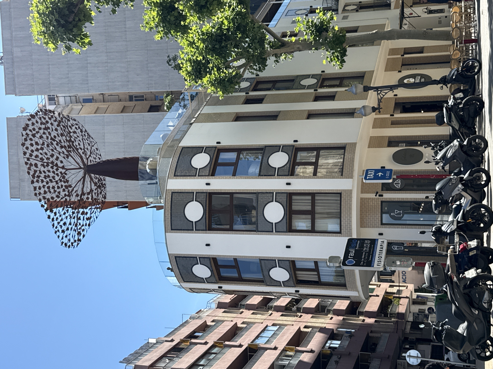This is a daytime street scene in Dénia, Spain. A building with a curved facade, featuring white panels and brown window frames, is centered. The building has a sign for "raul fisio," advertising physiotherapy services. A "TAXI" sign is also visible. Modern-style black motor scooters are parked on the street. A metal tree sculpture with leaf-like cutouts stands on the left. Buildings of varying heights with light and dark brown facades are in the background, against a clear blue sky. Branches of a tree obscure parts of the upper section of the frame.