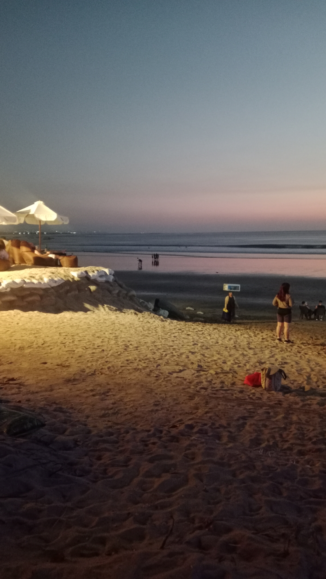 This is a medium shot, outdoor, vertical photograph of Kuta Beach in Kuta, Indonesia, taken at dusk. The sky is a gradient of deep blue at the top, transitioning to a soft pink and orange hue near the horizon. The ocean is calm with gentle waves rolling towards the shore. In the foreground, a sandy beach is visible with scattered footprints and debris. Two white beach umbrellas are set up on a raised sand platform on the left side of the image, with seating arrangements underneath.
Further back on the beach, several people are visible. One person is walking with what appears to be a flat, rectangular object balanced on their head, possibly a tray or a sign. To the right, a woman is standing, facing away from the camera, wearing shorts and a top. Near her, a backpack and a red bag are placed on the sand. In the distance, closer to the water, a group of people are standing together, and several other small figures are dispersed along the shoreline and in the shallow water. Lights from the town are faintly visible in the far distance on the horizon. The overall lighting suggests it is twilight, with artificial lights illuminating the foreground sand.