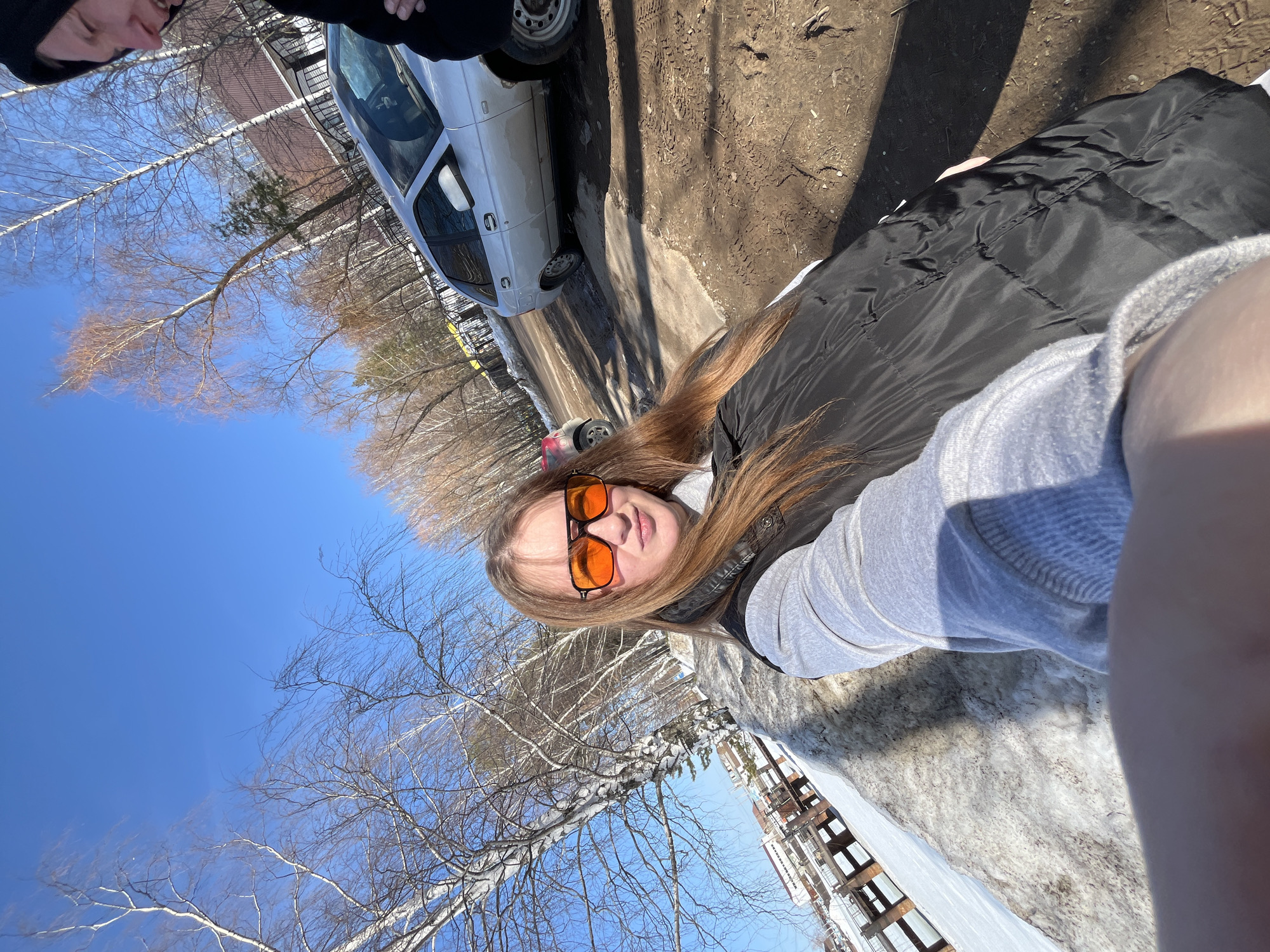 The image depicts an outdoor scene in Chaikovsky, Russia, characterized by bright sunlight, a clear blue sky, and evidence of melting snow.
In the foreground, a young female, estimated to be in her twenties, with long, light brown hair, is centrally positioned. She wears large, orange-tinted aviator-style sunglasses, a light grey long-sleeved shirt, and a black puffer vest. She is looking directly at the camera, holding the device with her right arm extended, suggesting a selfie. Her shadow is visible on her chest and the ground.
To the far right of the frame, a portion of a second individual is visible, including the lower part of their face (nose, mouth, chin) in profile, wearing a dark hooded garment. This individual appears to be smiling or speaking, facing away from the primary subject.
The ground consists of reddish-brown mud and dirt, displaying visible tire tracks and uneven surfaces. Patches of melting, dirty snow are interspersed throughout the ground and along a raised embankment to the left.
A silver Lada 2112 or similar hatchback vehicle is parked on the dirt road behind the subjects, with its front end facing right. A smaller, red vehicle is partially visible further in the background, to the left of the silver car.
The background features numerous deciduous trees with bare branches, predominantly white-barked birches, lining what appears to be a road or path. Beyond these trees, several light-colored buildings with dark, possibly tiled, roofs are partially visible. To the far left, a significant expanse of flat, snow-covered terrain, likely a frozen body of water, extends into the distance, bordered by a dark, multi-level wooden or concrete structure, possibly a pier or embankment support.