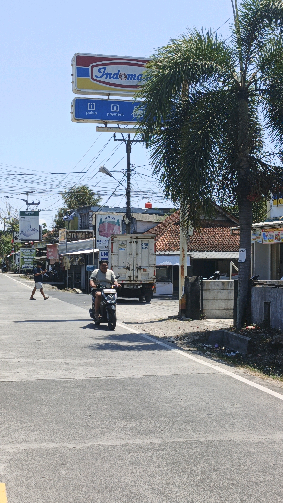 In Cibenda, Indonesia, on a sunny day, a street scene unfolds with commercial establishments and active traffic. Dominating the skyline is a prominent Indomaret sign, a popular convenience store chain, accompanied by smaller signs indicating "pulsa" (phone credit) and "payment" services. A tall palm tree with lush green fronds partially obscures the view. In the foreground, a man rides a black motorcycle, wearing a light-colored t-shirt and dark shorts. He is traveling on an asphalt road marked with white lines. To the left of the road, a pedestrian in a dark shirt and light shorts walks across the street. Behind them, various shops and buildings line the road, including a white truck with the license plate "CRB 605" parked near a building with a red tile roof. Several power lines are visible overhead, crisscrossing the sky. The environment appears to be a bustling local area with signs of commerce and everyday life.