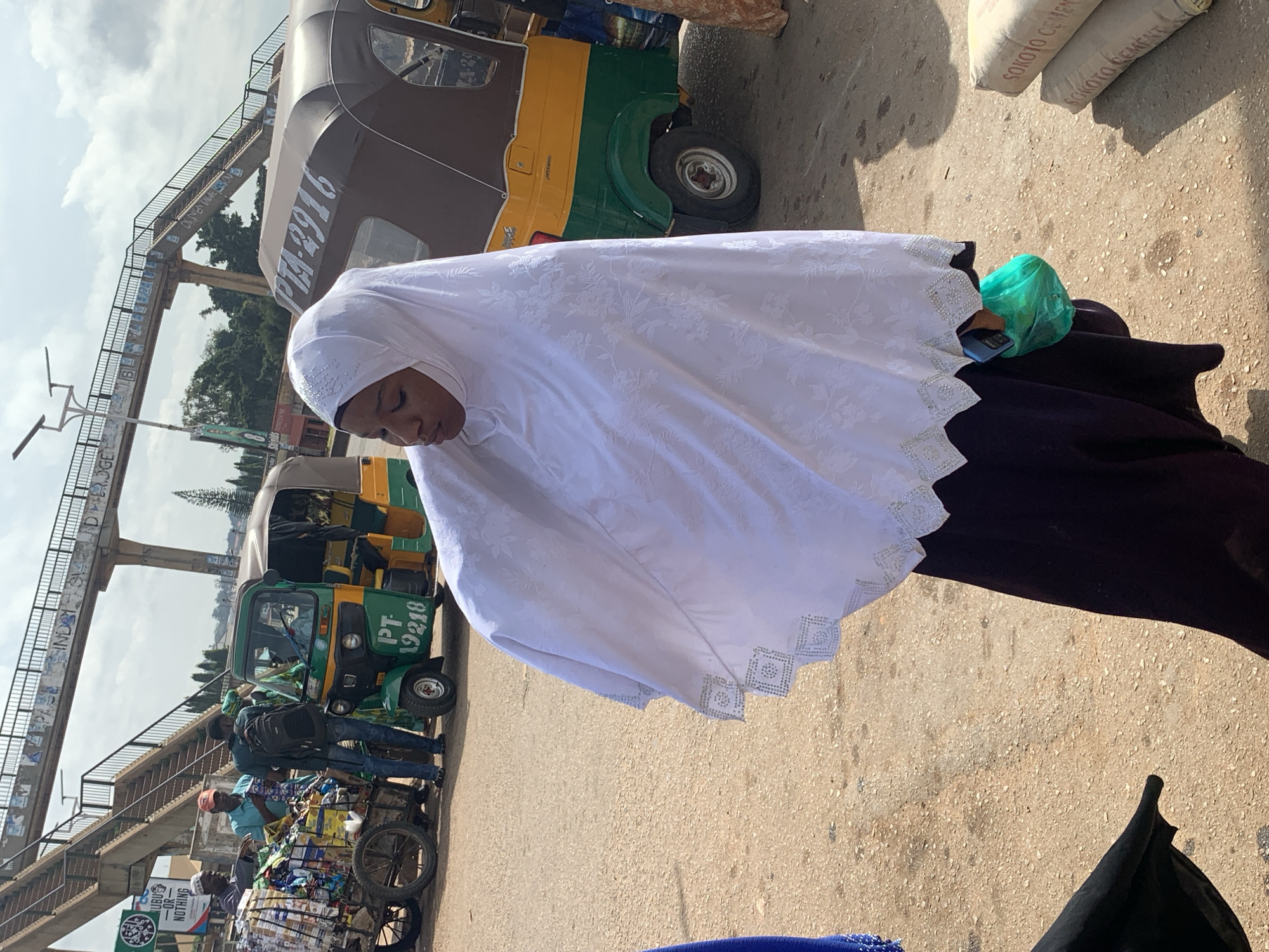 In Gura Top, Nigeria, a young woman in a white hijab and dark purple dress stands on a dirt road. She holds a green plastic bag and her head is bowed. Behind her, a yellow and green auto-rickshaw is parked, displaying the license plate PTA-2916. Further in the background, a pedestrian overpass spans the road, with signs visible underneath. To the left, a cart filled with various goods is being attended to by several men. Bags of "Sokoto Cement" are stacked on the right side of the image. The scene appears to be a busy marketplace area with elements of urban infrastructure.