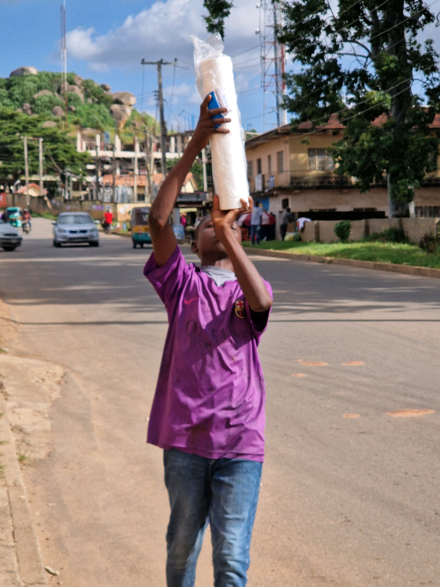 A young African male, wearing a purple t-shirt and blue jeans, stands in the middle of a paved road in Jos, Nigeria. He is holding a large, white, cylindrical object wrapped in plastic above his head with both hands, looking upwards. The object appears to be a roll of something, possibly paper towels or similar material. The road is lined with trees and buildings on either side. In the background, there are cars, motorcycles, and people. The sky is blue with scattered clouds. There are also large radio towers visible in the distance, suggesting the location is within a populated area of the city.