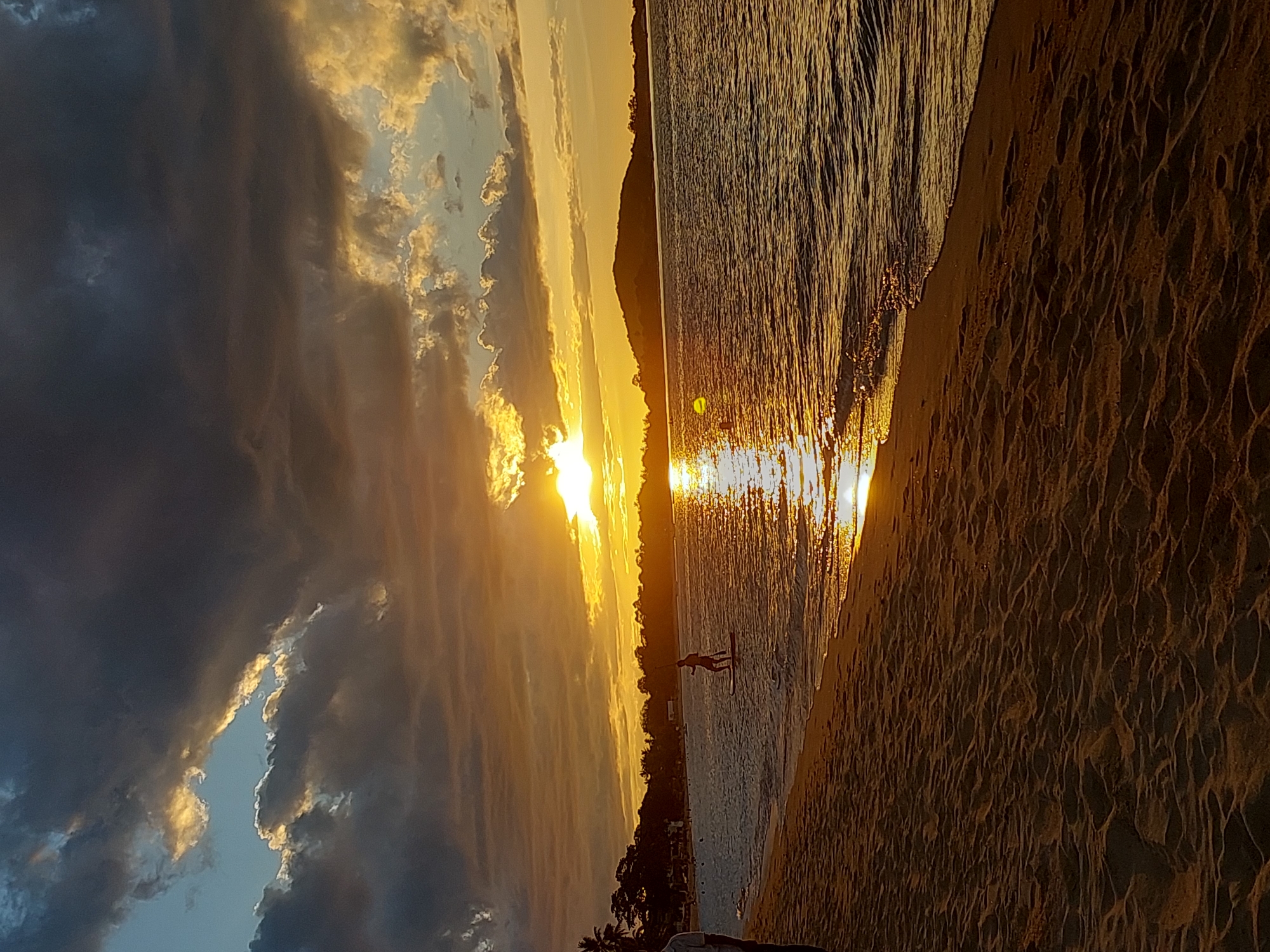 This is a vertical, wide-angle, eye-level shot of a beach at sunset. The sun is low on the horizon, casting a warm, golden light across the sky and water. Dark, dramatic clouds fill the left side of the frame, contrasting with the bright, illuminated sky on the right. The ocean is calm, with ripples reflecting the sunset's glow.
A person is visible in the water, standing on what appears to be a paddleboard. They are a silhouette against the bright light, their posture suggesting they are either paddling or posing. The beach in the foreground is sandy, with textured patterns created by the receding tide or wind. Trees and a distant shoreline are visible across the water.
The time of day is clearly sunset, indicated by the low angle of the sun and the warm colors in the sky. The weather appears to be fair, with a mix of clouds and clear sky, creating a dramatic visual effect. The location is a tropical beach, with palm trees visible. The overall emotion conveyed is peaceful and serene, with a touch of awe inspired by the natural beauty of the sunset. There is no visible text in the image.