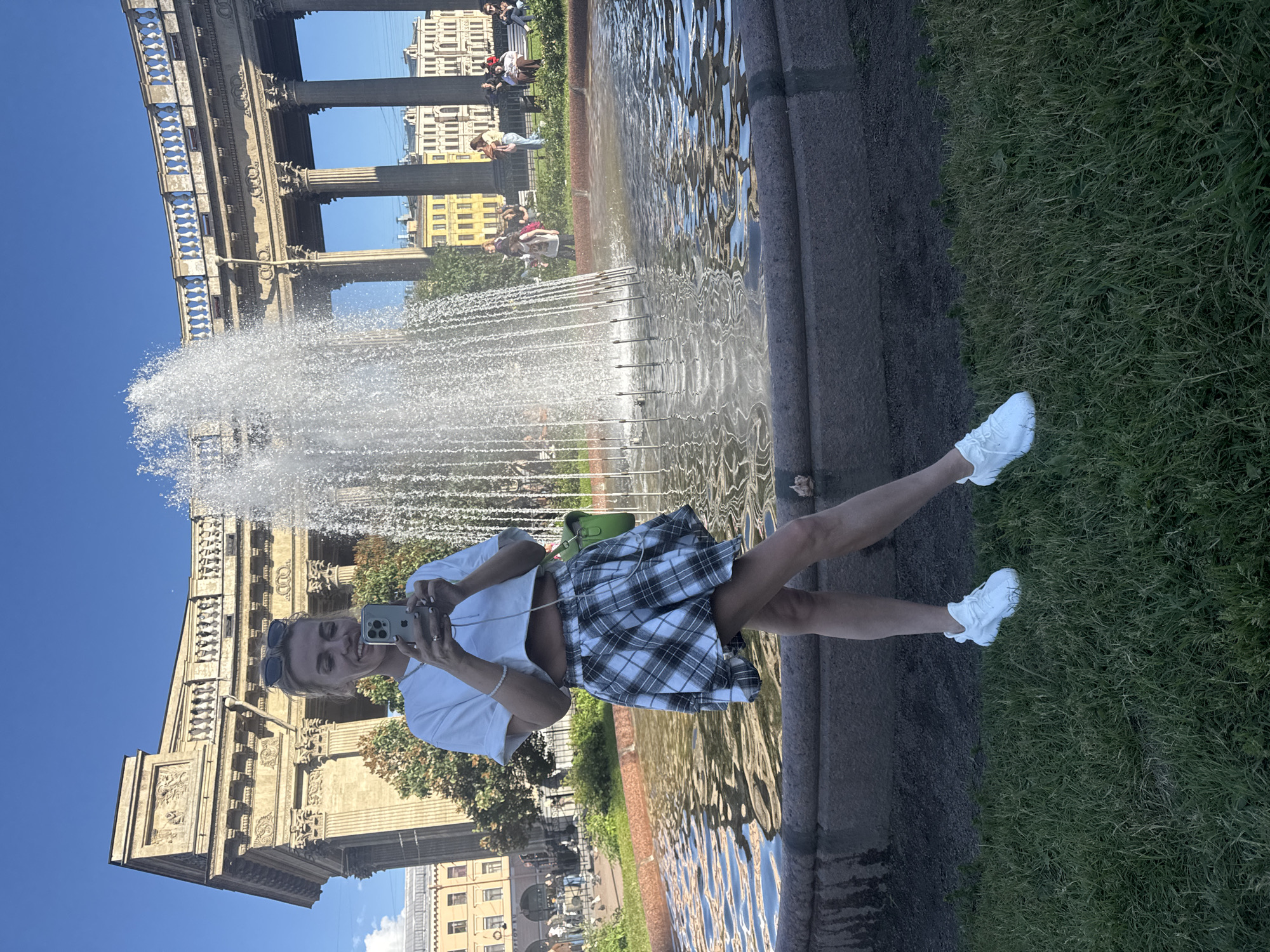 A young woman is taking a selfie in front of a large, ornate fountain with a grand architectural building in the background. She is wearing a white crop top, a black and white plaid skirt, and white sneakers. She has sunglasses perched on her head and is smiling at the camera while holding up her phone. A green purse is slung over her shoulder. The fountain is spraying water, creating a misty effect. The building behind her appears to be made of stone with classical architectural elements, including columns and decorative carvings. The sky is a clear, bright blue, suggesting a sunny day, likely daytime. The ground around the fountain is paved, with grass visible to the right. People can be seen in the background, some sitting on benches and others walking, indicating a public space. The overall impression is one of a tourist enjoying a sunny day in a historically significant location.