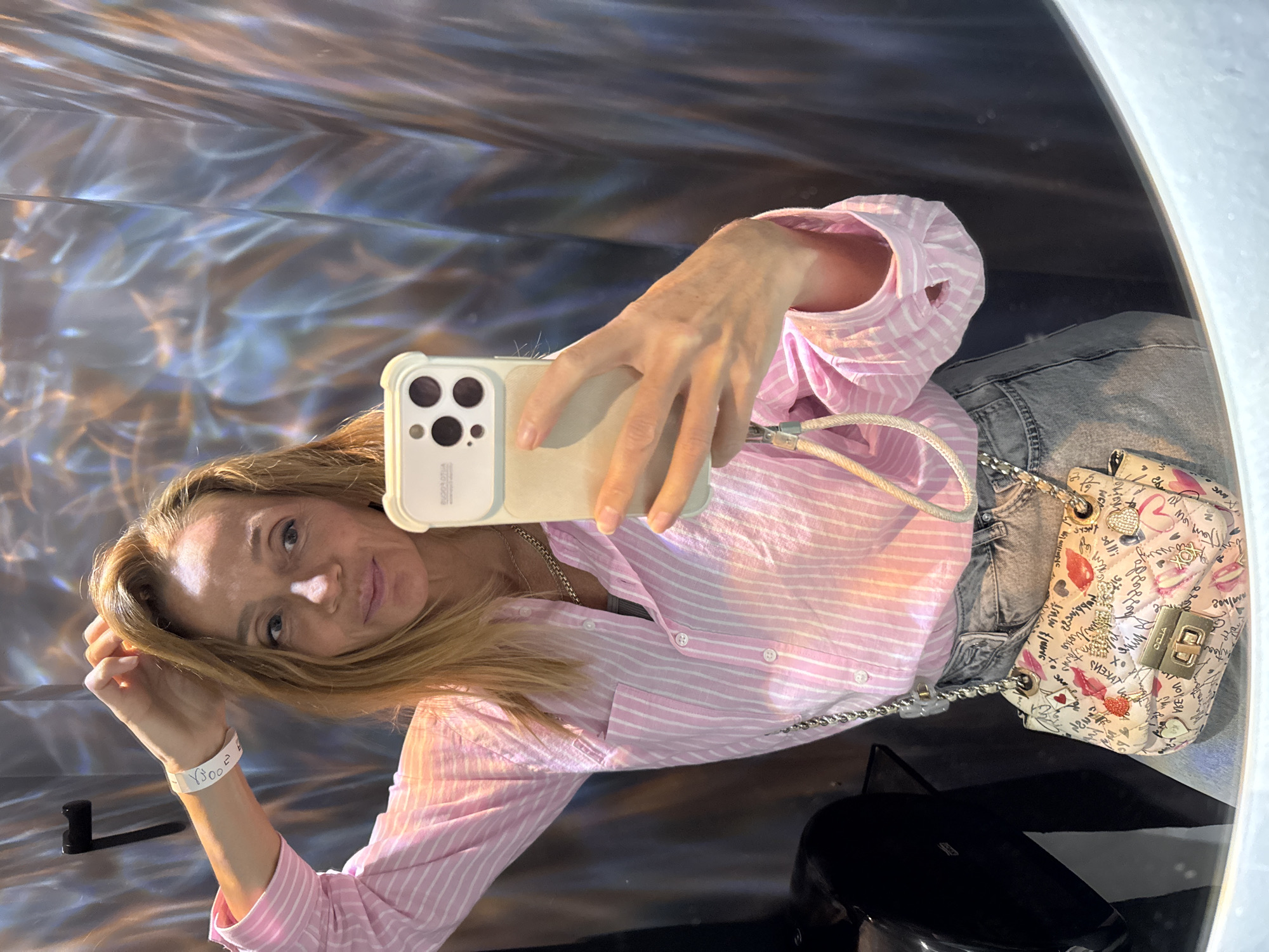 A blonde-haired woman takes a selfie in what appears to be an art installation or a room with reflective surfaces. She is wearing a pink and white striped button-down shirt and jeans. A white handbag with a chain strap rests beside her. The environment consists of swirling patterns, possibly caused by light reflecting off the surfaces. A black, shiny object is also visible. No explicit location information from Saint Petersburg, Russia, can be discerned from the image.