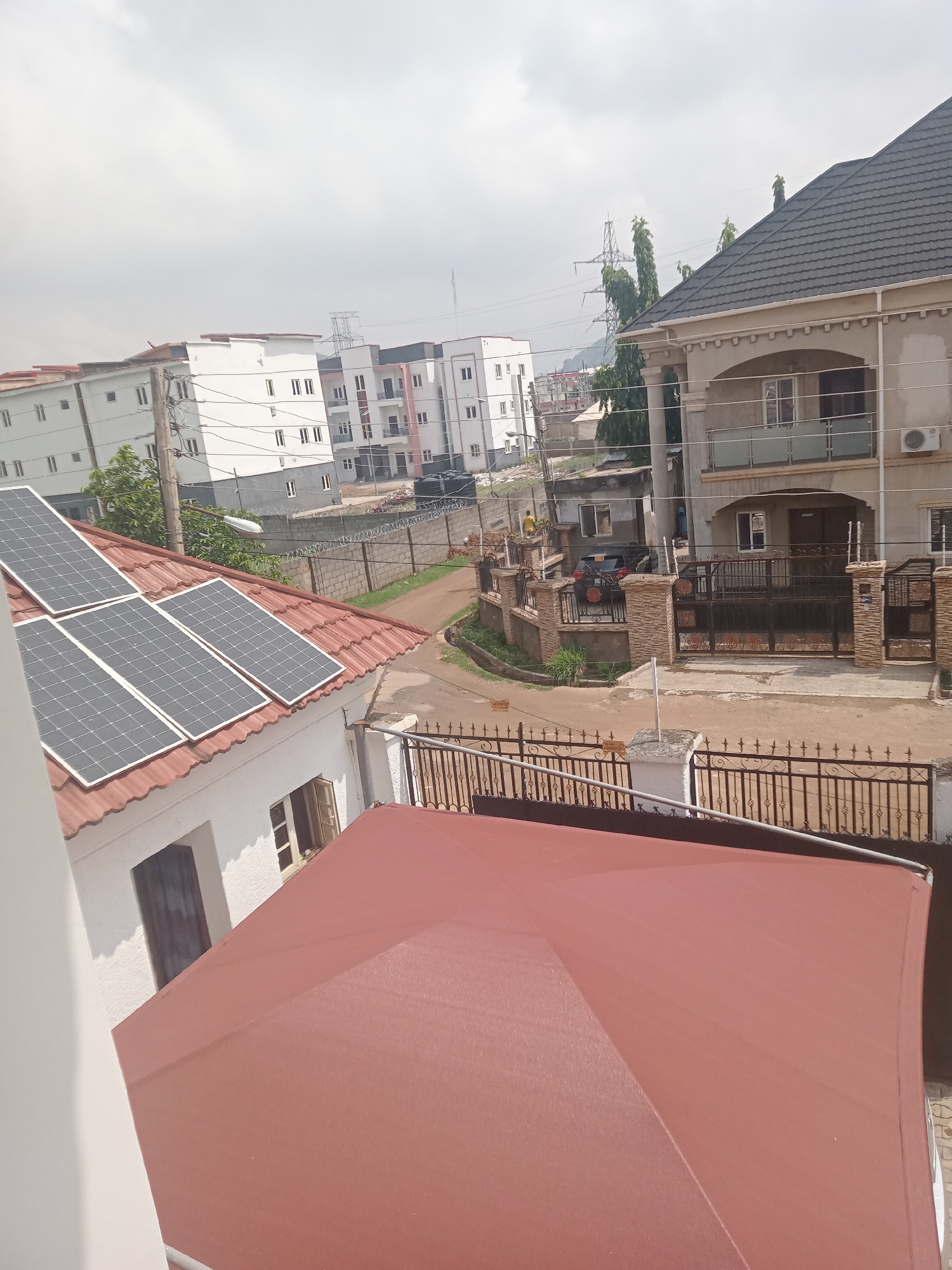The image shows a residential area in Gwarinpa, Nigeria. Several multi-story buildings are visible, along with individual houses with gated entrances. Solar panels are installed on the roof of one house. Power lines and utility poles are present. A person in a yellow shirt is visible in the distance. Vehicles are parked on the street. The overall environment suggests a middle-class neighborhood with a mix of building styles.