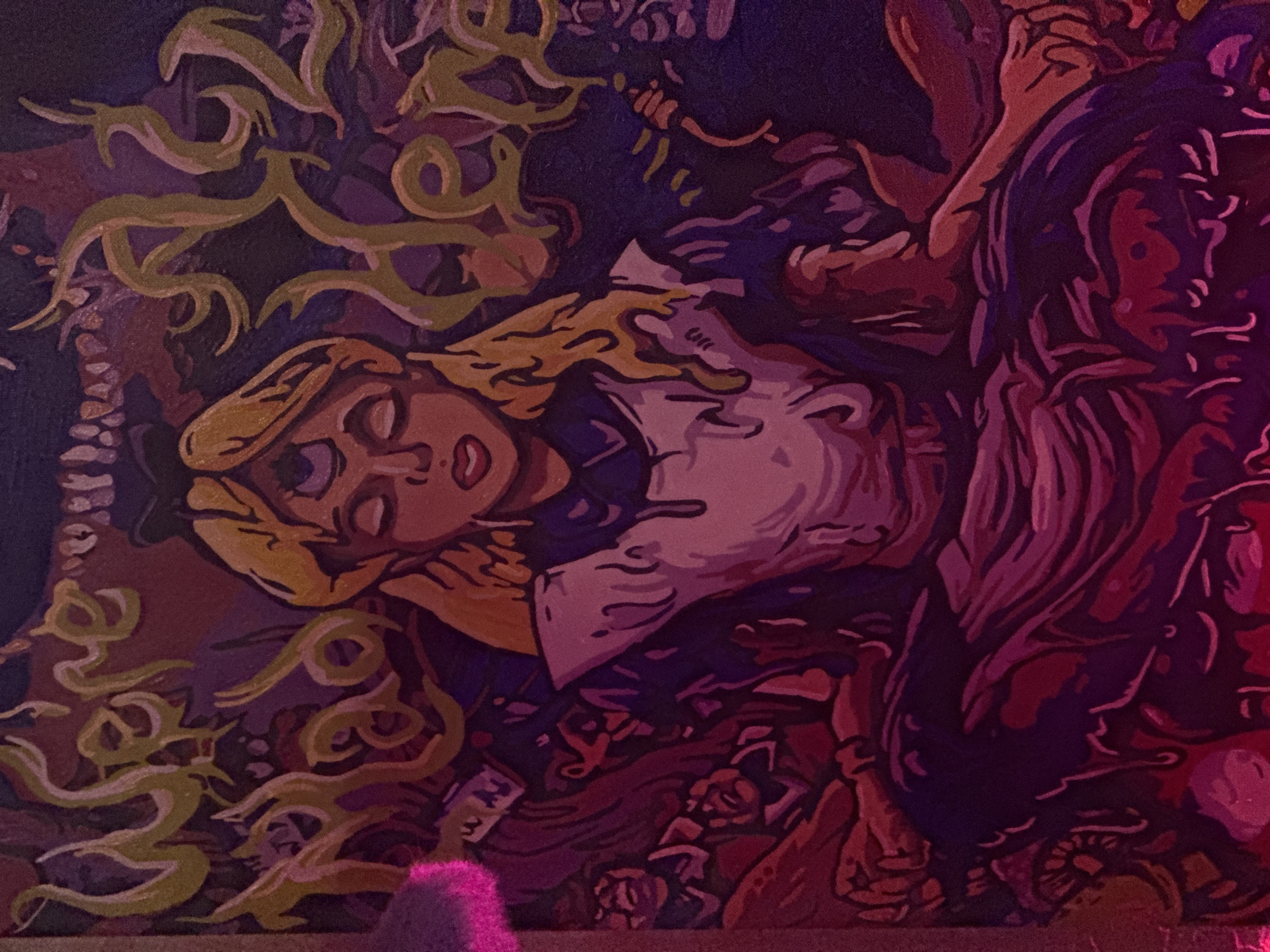 The image is a close-up, slightly blurry painting of a stylized Alice from Alice in Wonderland. She is depicted with three eyes on her forehead, closed. Her hair is blonde and wavy, and she is wearing her iconic blue and white dress. Around her head and shoulders, swirling green text reads "We're all mad here." The background is dark and chaotic, with abstract shapes and figures rendered in shades of purple, red, and pink. The overall impression is surreal and unsettling, with a focus on the theme of madness. The painting is illuminated by a pinkish light source, which casts a glow on the left side of the image and also seems to be the dominant light source in the scene. There are no discernible city elements or specific locations within the image.