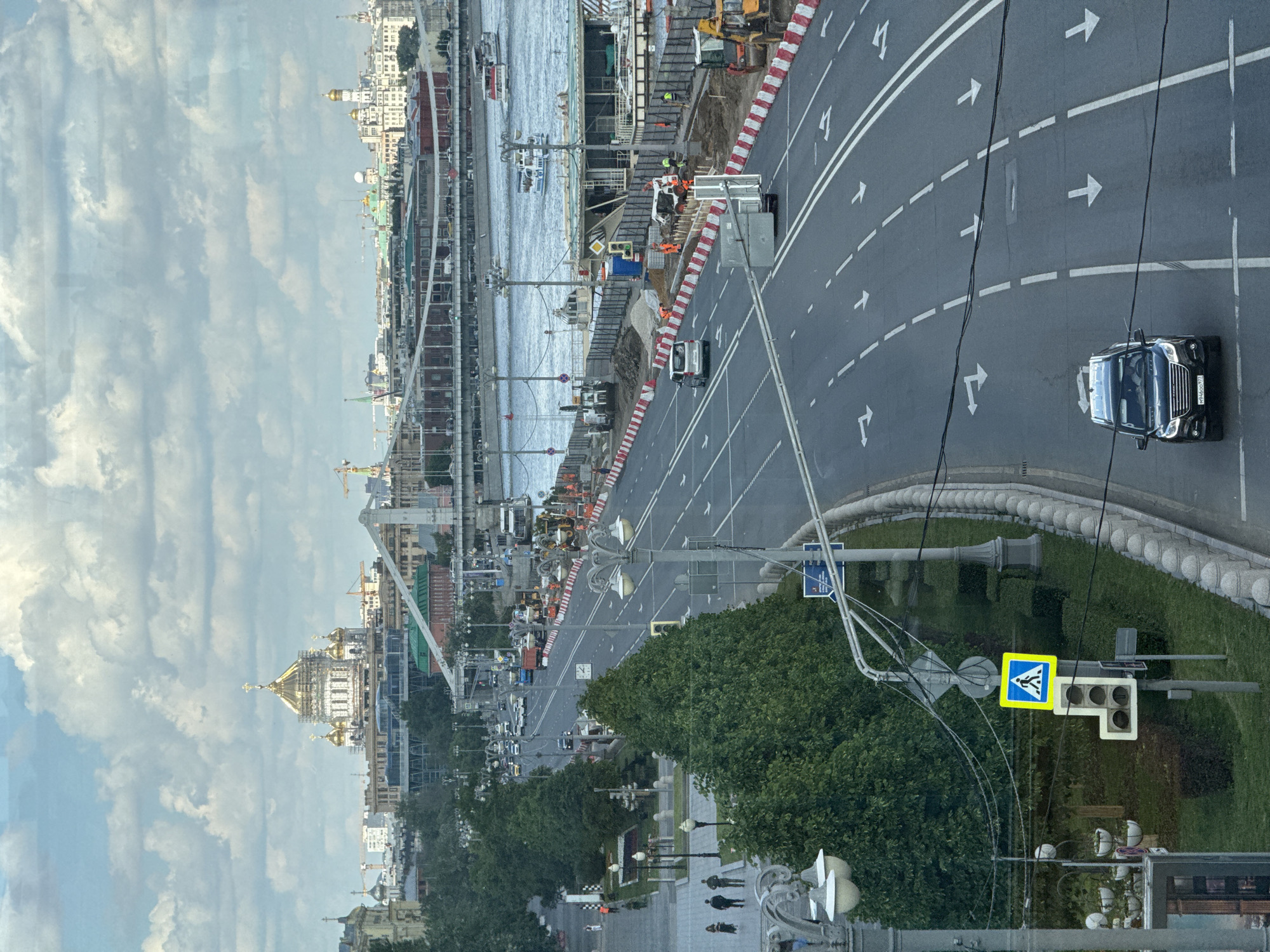 The image depicts a wide panoramic view of an urban landscape along a river, likely captured from a building overlooking the scene.
In the foreground, a multi-lane asphalt road runs parallel to the river. White painted arrows on the road indicate traffic flow, mostly straight ahead, with some turning lanes. A black car, possibly an SUV or van, is prominently featured in the rightmost lane. Its license plate, "M880OP799", is visible. To the right of the road is a green area with trees, grass, and a paved walkway where several pedestrians are seen. Streetlights and decorative lampposts line the walkway. A yellow diamond-shaped sign indicating a pedestrian crosswalk is visible on a pole.
The middle ground shows extensive construction work along the riverbank. Red and white striped barriers delineate the construction zone. Several construction vehicles, including excavators and trucks, are present, along with workers in high-visibility orange vests. Boats are visible on the river, including a white passenger vessel.
In the background, on the opposite side of the river, a dense cityscape stretches into the distance. A prominent feature is a large golden-domed church, possibly undergoing some renovation as scaffolding is visible around parts of it. Other buildings of various architectural styles are visible, some with cranes nearby, suggesting further development.
The sky is partly cloudy, with scattered white clouds against a lighter blue. The time of day appears to be daytime, with ample natural light. The overall scene suggests a busy city environment with ongoing infrastructure development and transportation activities on both land and water. The image seems to be taken through a window, as subtle reflections are visible on the left side.