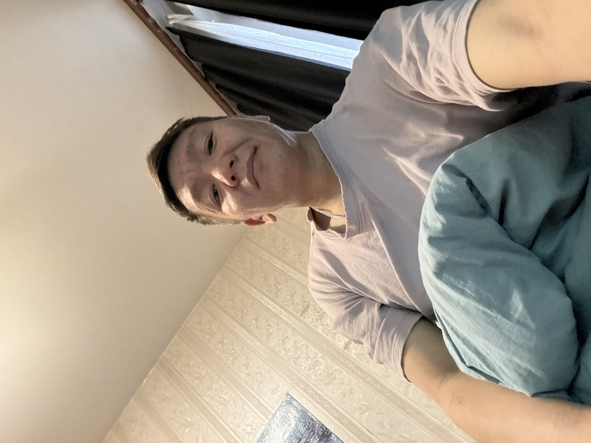 The image shows a low-angle, waist-up selfie of an East Asian male in a room. He is wearing a light purple t-shirt and a silver chain necklace. He is holding a light blue comforter or blanket, partially covering his torso and arms. The background features patterned wallpaper on the left and dark curtains with a white sheer curtain visible behind them on the right. The ceiling is visible at the top of the frame. The lighting suggests an indoor environment, possibly a bedroom. The location is identified as Khimki, Russia.