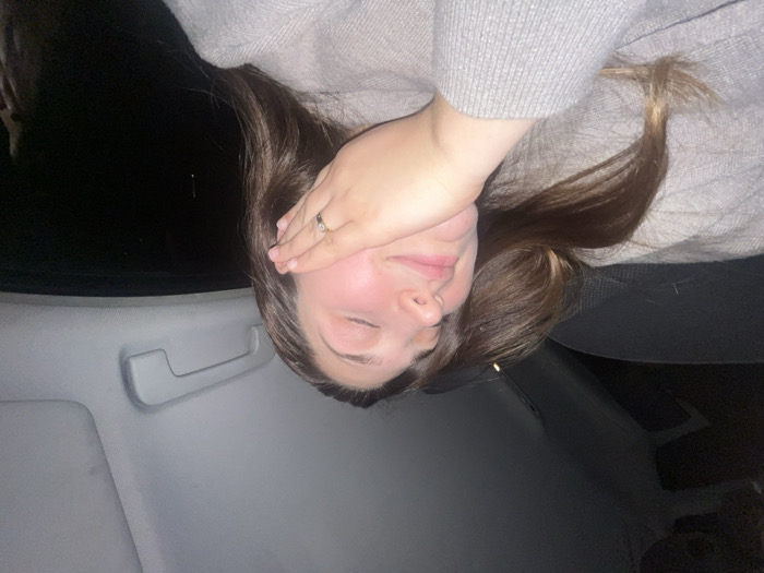 A woman with brunette hair reclines in a car seat, her eyes closed. She wears a light gray sweater and a ring on her left hand. Her hand shields her face from the light. The interior of the car is visible, including the gray car door and a section of the seat. Outside, the night is dark and obscures the background. The image is slightly rotated.