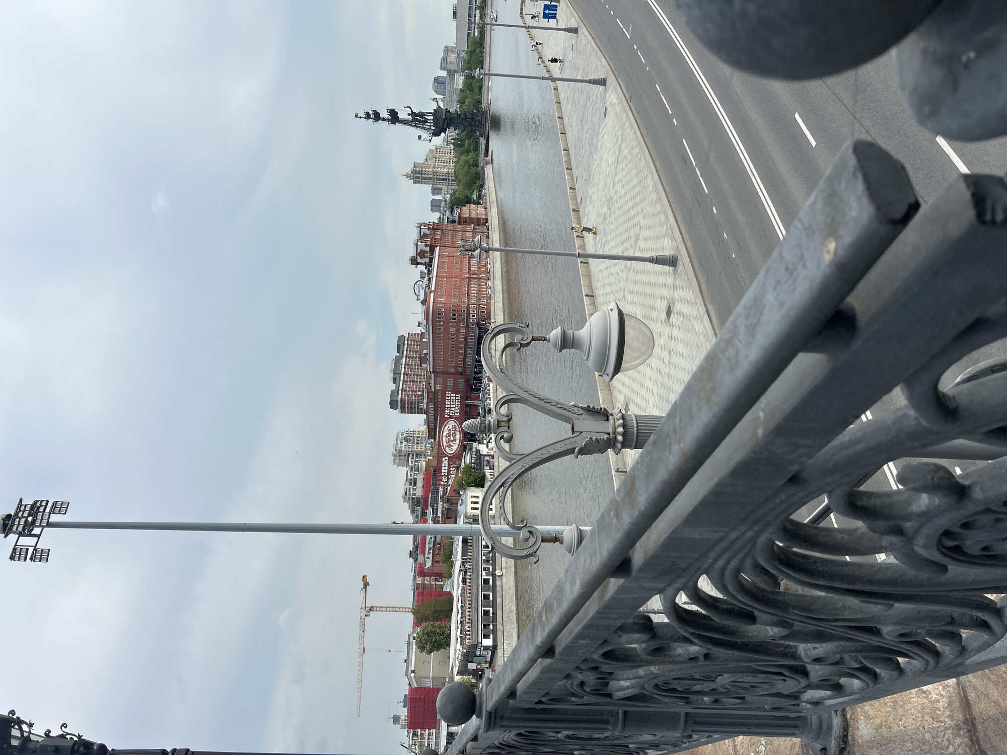 A slightly rotated, high-angle shot overlooking the Moskva River in Moscow, Russia. A decorative, wrought-iron bridge railing is in the foreground. Beyond the railing is a street with several white lines dividing the lanes. A lamp post is centered in the midground. The Moskva River is in the center of the frame, with buildings on both sides of the river. A large statue stands in the middle of the river on an island. In the background, more buildings can be seen, and the sky is overcast. To the left, a tall lamppost and a construction crane are visible against the sky.