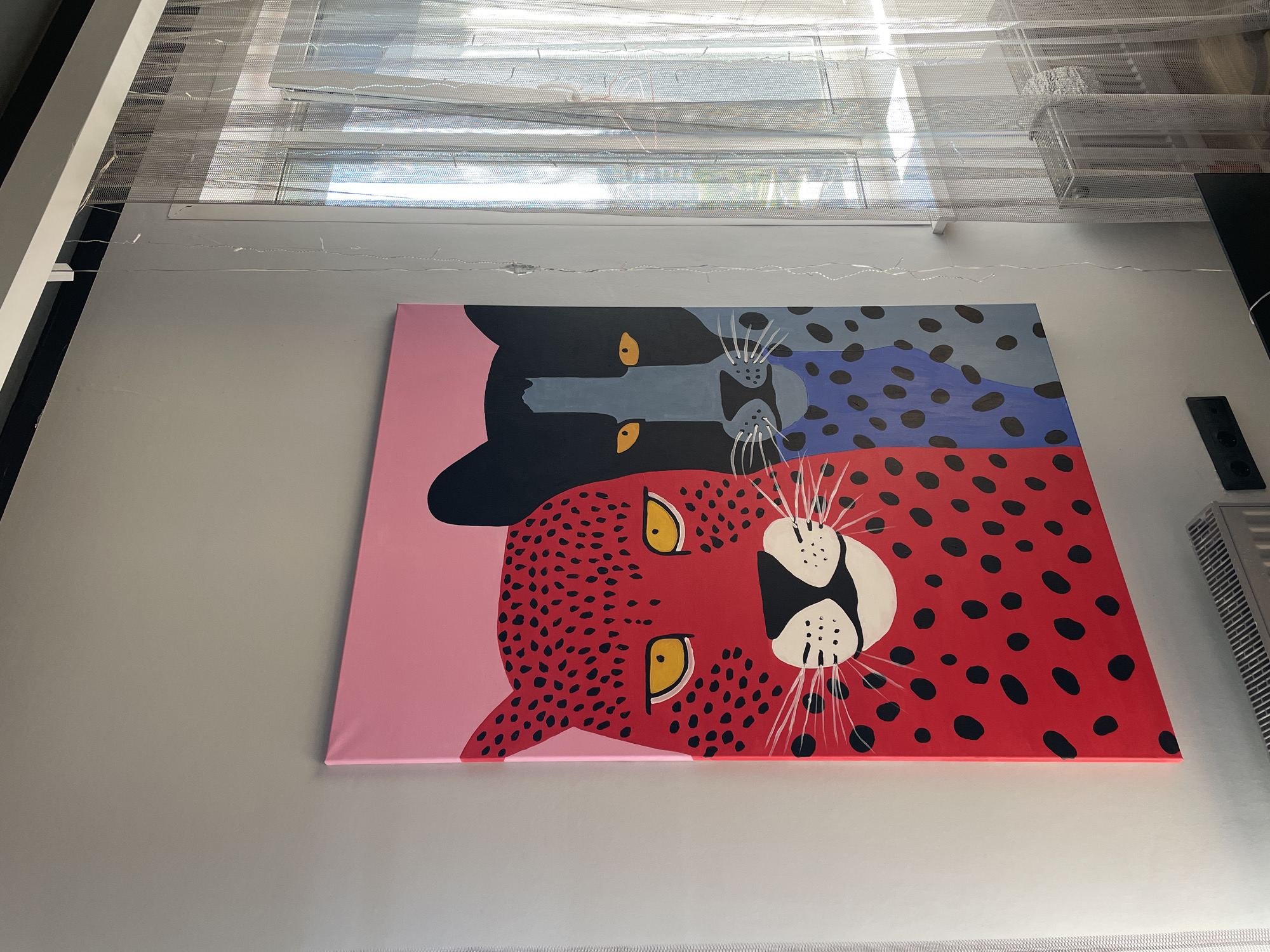 A close-up, eye-level shot shows a colorful painting of two leopards hung on a white wall beneath a window with partially drawn white blinds. The painting is divided in half vertically, with the top half featuring a black leopard with blue spots on a blue background, and the bottom half displaying a red leopard with black spots on a pink background. The heads of the leopards are oriented towards each other in the center of the painting. A small black electrical outlet and a white air vent are visible on the wall to the right of the painting. A string of fairy lights is loosely strung along the wall near the window. The image was taken in an indoor location, and is not associated with Krasnoyarsk, Russia.