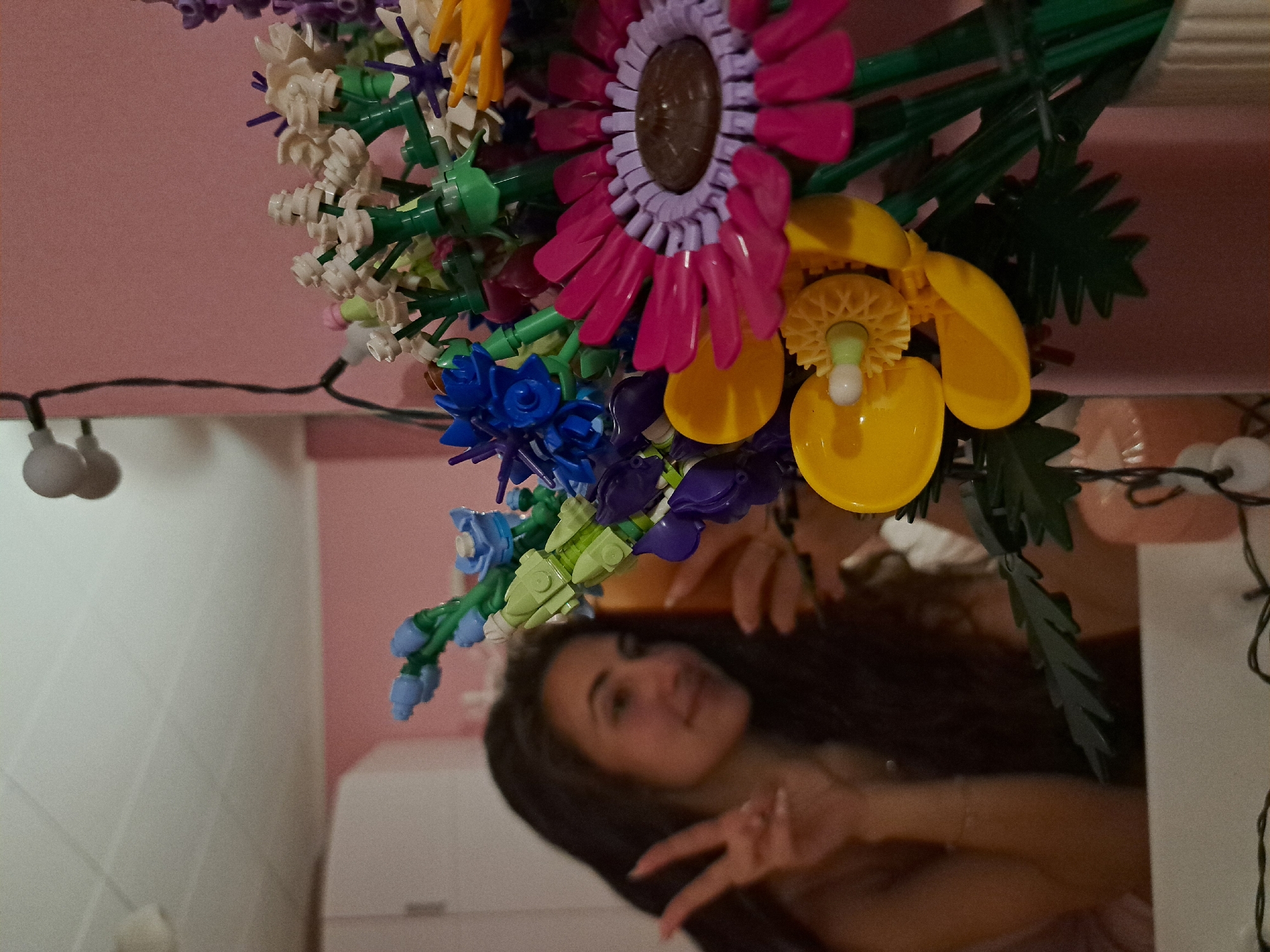 An overhead shot captured from below showcases a vibrant bouquet of artificial flowers, crafted from colorful building blocks like Lego or K'nex, set against a light pink background. The bouquet features a variety of flower types and colors, including a prominent central flower with bright pink petals and a purple center, yellow flowers with intricate centers, white/cream flowers, and smaller blue and purple floral elements. Green stems and leaf pieces are interspersed throughout the arrangement, creating a full and lively display.
Below the bouquet, a clear reflection reveals a young woman looking up at the flowers. Her long, dark hair frames her face, which appears slightly out of focus. She is seen smiling softly and has one hand raised, making a peace sign with her fingers. The reflection also shows parts of an indoor setting, including light-colored walls or furniture. String lights with small, round, white bulbs are visible both directly in the foreground, hanging below the flowers, and reflected in the lower part of the image, suggesting a cozy or decorated environment. The image is composed in a way that suggests the camera was held below the flowers, capturing both the actual bouquet and the reflection of the person holding or observing it.