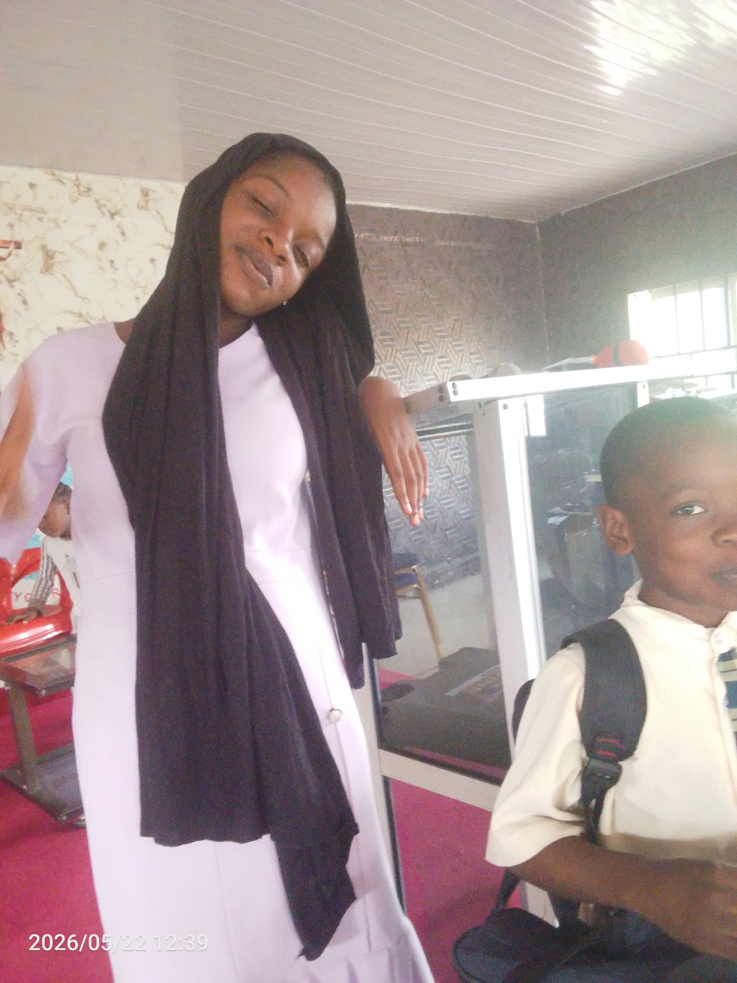 The image shows a woman and a young boy indoors. The woman is wearing a long-sleeved, light-colored dress and a black headscarf. She is leaning on a white counter or display case. The boy is wearing a light-colored shirt with a backpack. He is standing to the right of the woman. The room has a patterned wall and a window. The date "2026/05/22 12:39" is printed at the bottom left of the image. The location is Lokoja, Nigeria.