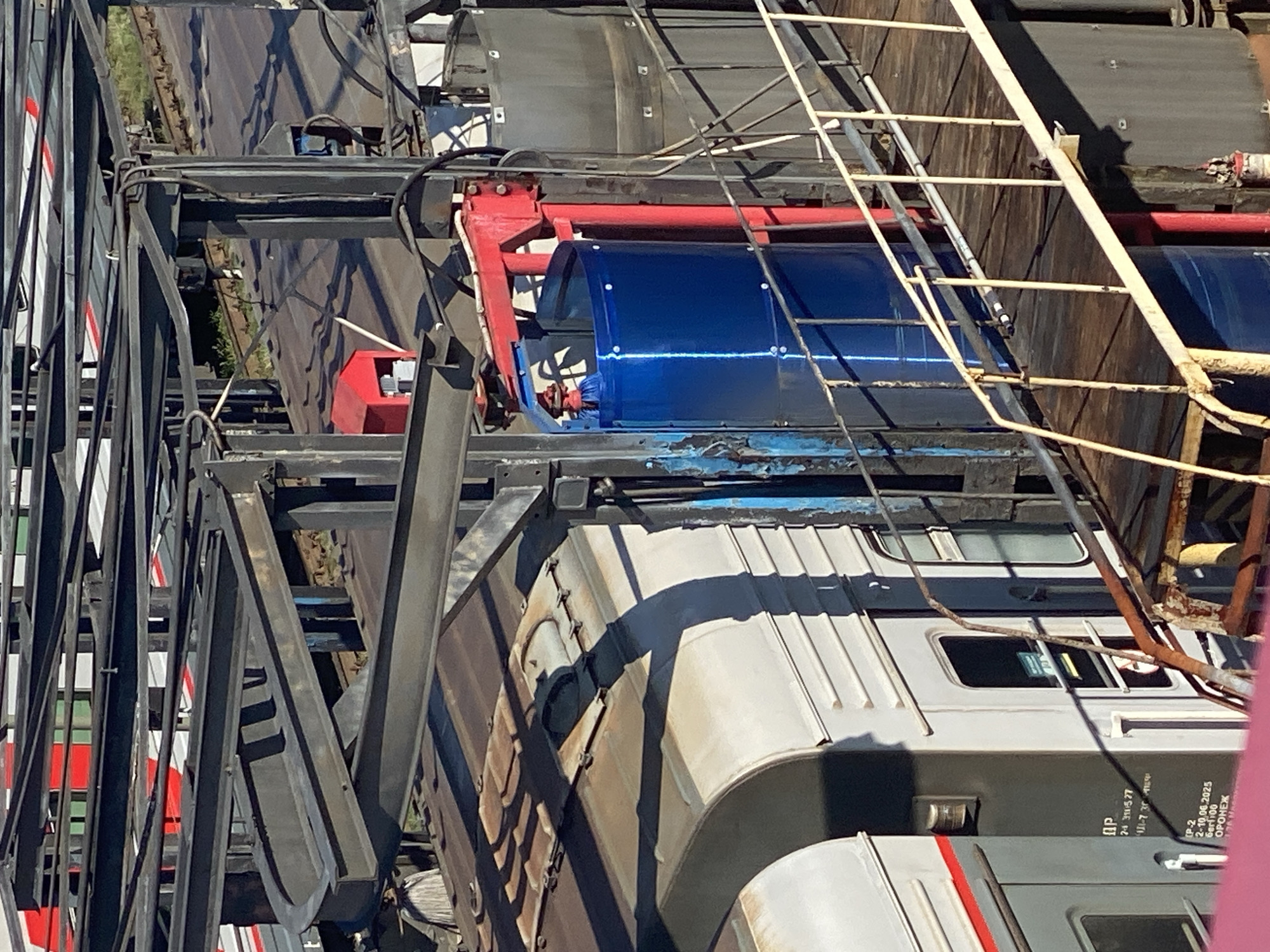 The image shows an outdoor industrial area, likely a train yard or depot, in Novokuznetsk, Russia. Prominent structures include overhead metal framework, possibly for an electric train line or a loading mechanism, and large cylindrical tanks. A train car, with a grey body and red and white detailing, is visible on the left. A bright blue cylindrical object, possibly a container or part of machinery, is positioned within a red metal frame structure, suggesting an active industrial process. A yellow metal walkway or staircase with railings runs across the lower right portion of the image, leading towards the machinery and tanks. The environment appears to be a sunny day, casting distinct shadows on the train and the ground.