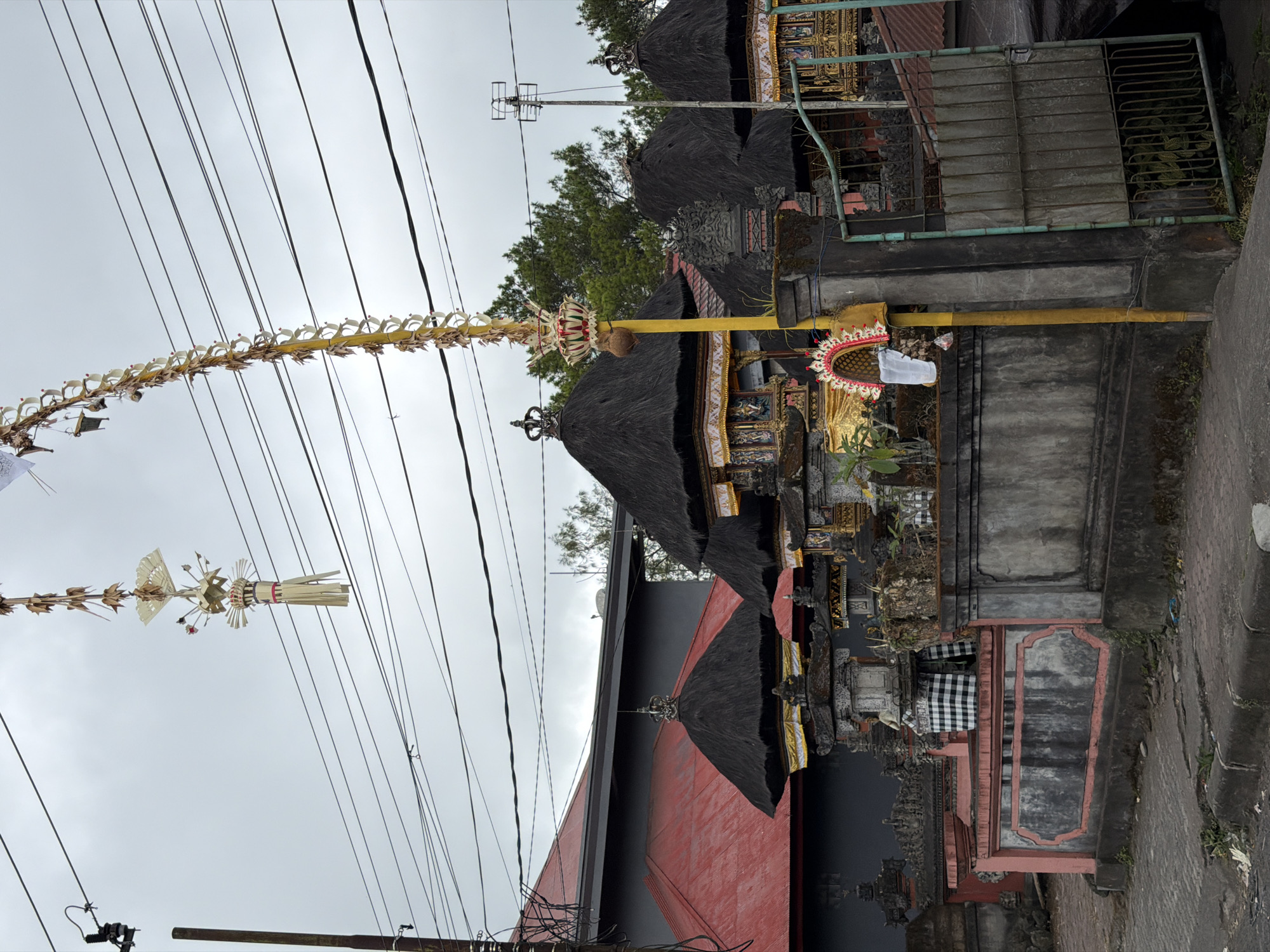 The image captures a street view in Kintamani, Indonesia, characterized by traditional Balinese architecture and overhead electrical wires. In the foreground, a low concrete wall with a pink trim borders a paved walkway. Beyond the wall, several structures with thatched roofs, characteristic of Balinese temples or homes, are visible. These buildings are adorned with ornate decorative elements, including intricate carvings and golden accents.
A tall, yellow pole stands prominently in the middle ground, decorated with a traditional Balinese offering known as a "penjor," which is a curved bamboo pole adorned with woven leaves and flowers. Hanging from a nearby overhead wire is another decorative element, possibly a smaller offering or ornament, made of dried leaves and woven material. The sky is overcast, suggesting a cloudy day. In the background, lush green trees are visible, hinting at a more rural or natural setting surrounding the built environment. An open green metal gate is situated on the right side of the image, revealing glimpses of more structures behind it. The overall scene conveys a sense of local culture and spiritual practices within an urban or semi-urban landscape.