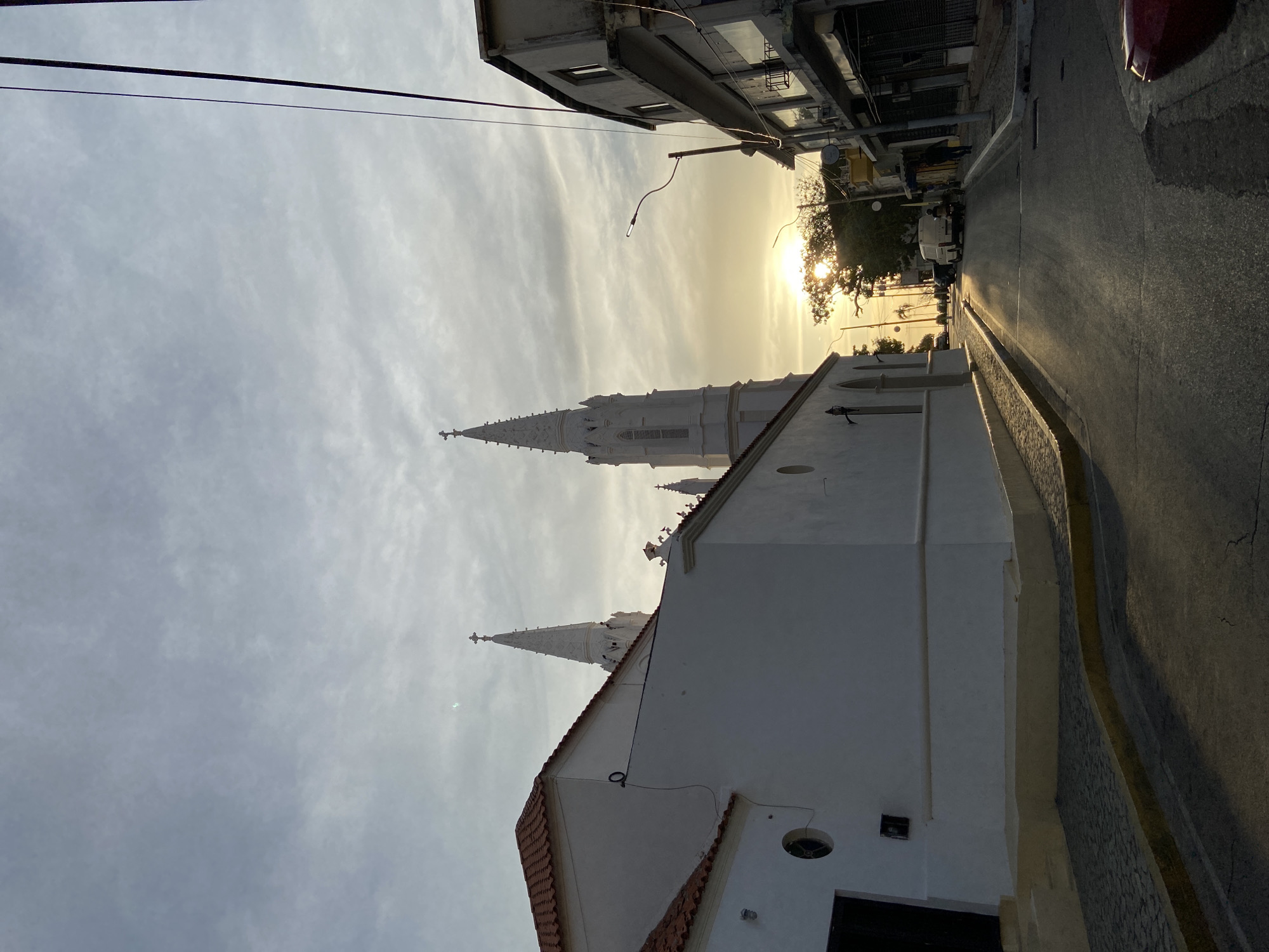 The image shows a street scene in Juan Griego, Venezuela, with a view toward the setting sun. A white building with a red-tiled roof and two prominent white steeple towers, likely a church, dominates the foreground on the left. A street runs along the right side of the image, marked by a yellow curb. Buildings line the street, partially obscured by the glare of the sun. A few vehicles are parked further down the street. Power lines stretch across the sky. The sky itself is overcast with light clouds.